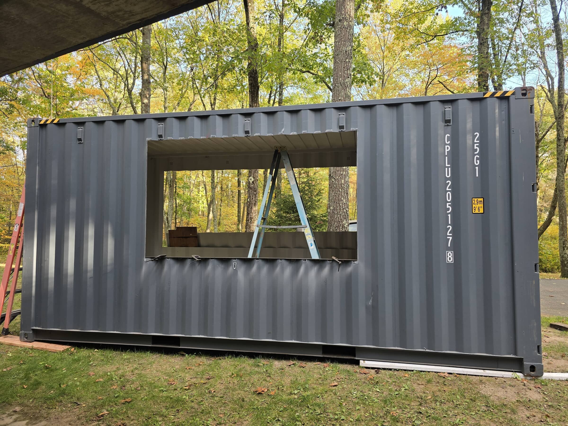 Construction site with a gray shipping container with a window opening, a ladder inside, surrounded by trees with fall foliage.