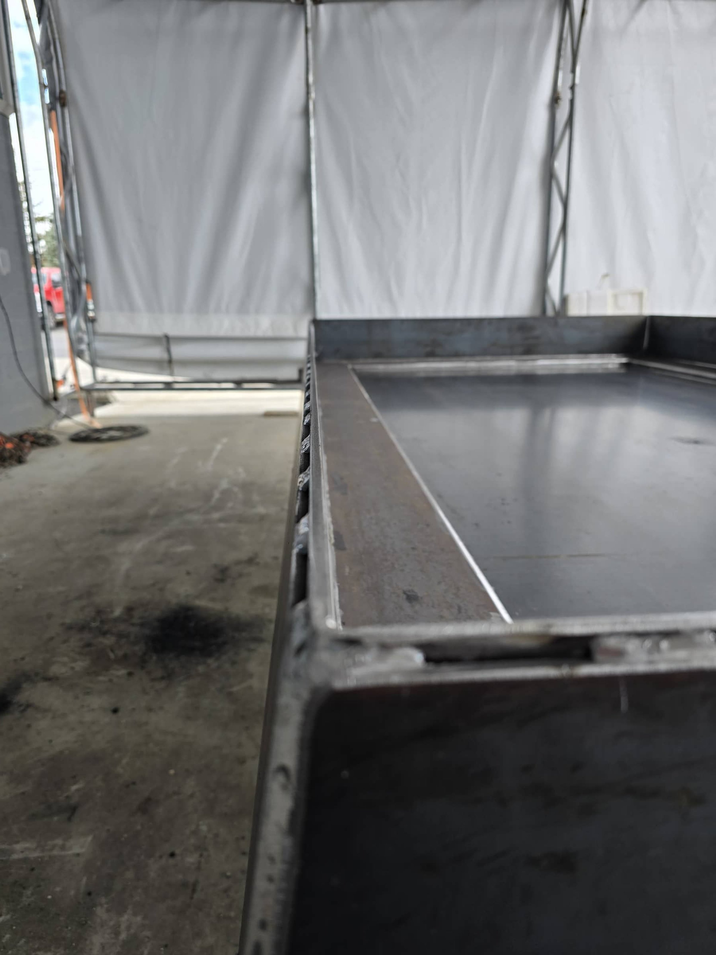 Close-up of an empty stainless steel industrial sink or worktable in a garage or workshop, with a white curtain or tarp in the background.