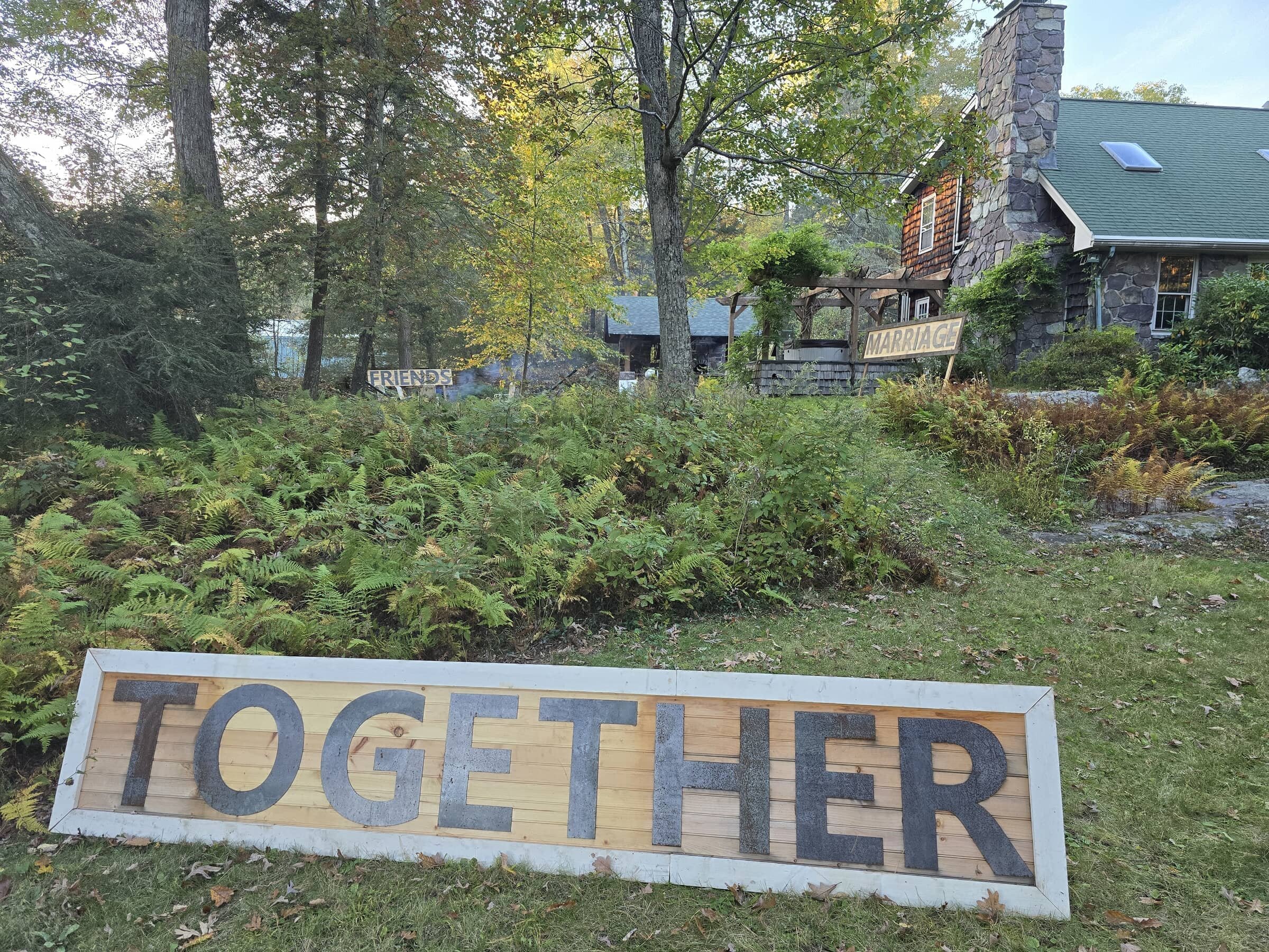 Large wooden sign with the word 'TOGETHER' in black letters, placed on the grassy ground. In the background, there are smaller signs reading 'FRIENDS' and 'MARRIAGE' near a stone house surrounded by trees and greenery.