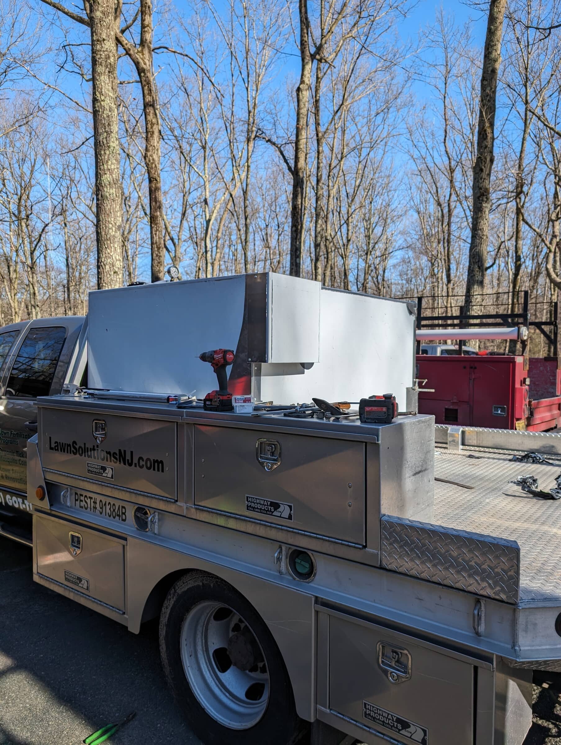 A utility truck with a metal workbench attached, containing a cordless drill, tools, and batteries, parked in a wooded area with leafless trees and a blue sky.