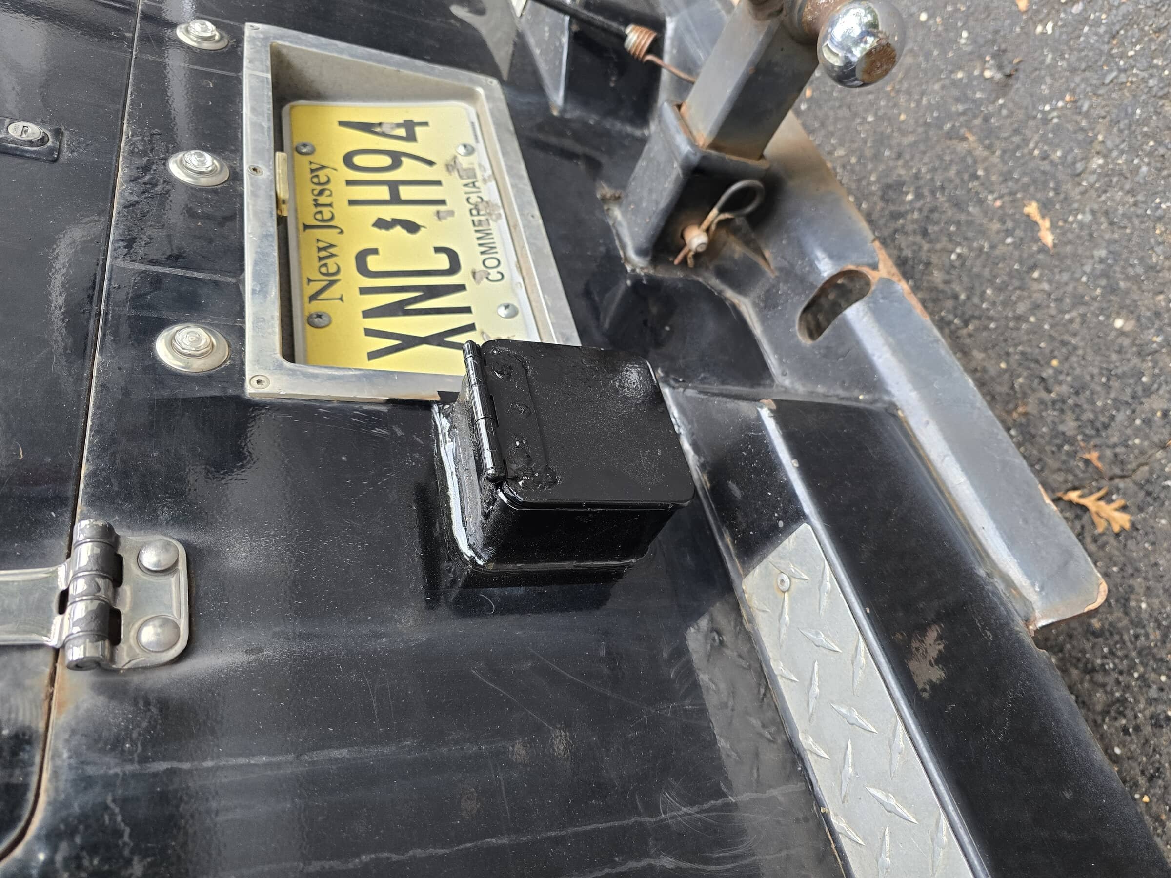 Close-up of a trailer hitch with a New Jersey license plate reading 'XNC 464' attached to the front, and a black electrical connection box nearby, on a paved surface with scattered leaves.