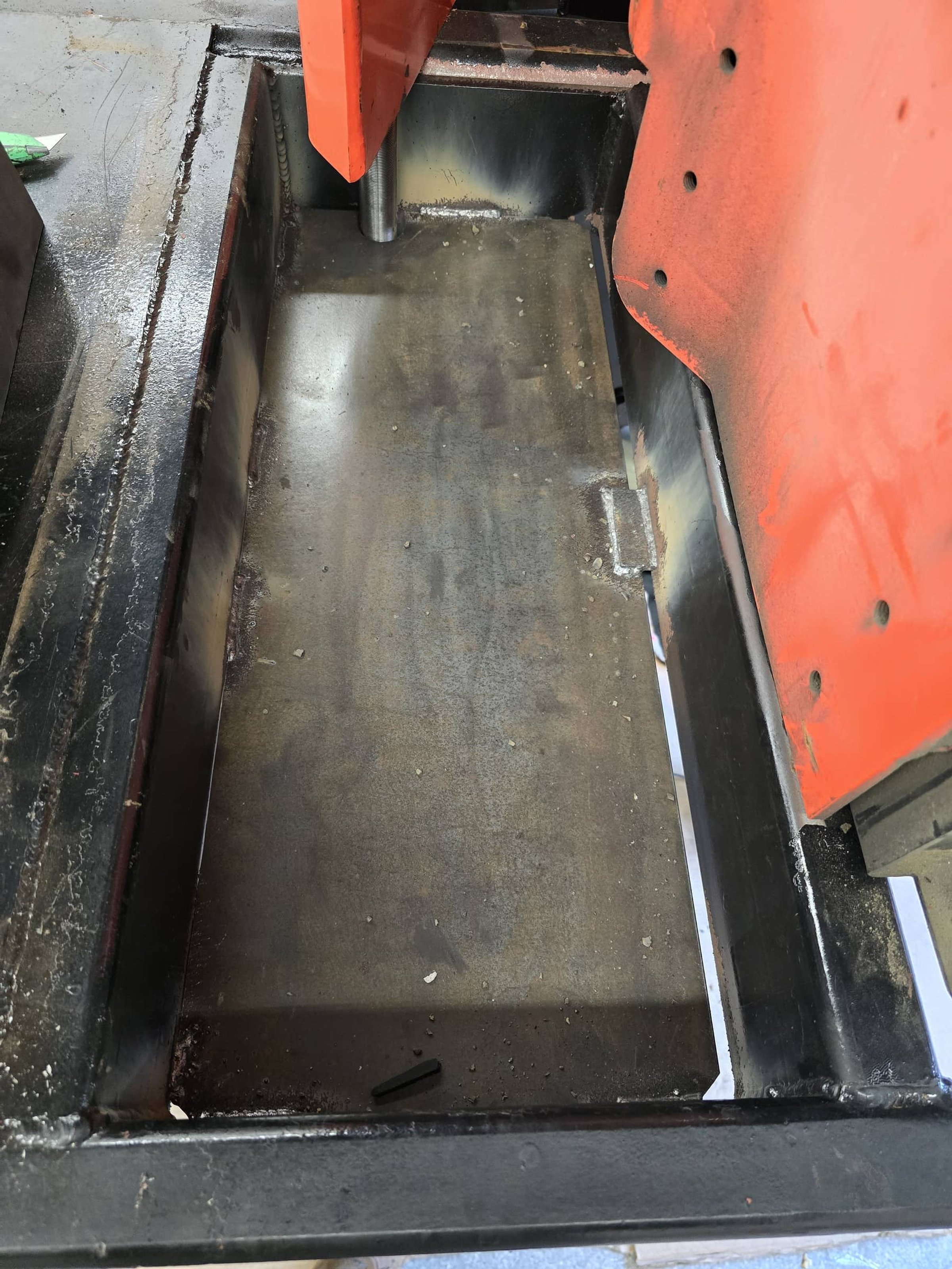 Interior of a metal workspace with a black welded plate, showing signs of use and grime, and part of an orange machine or tool in the upper right corner.
