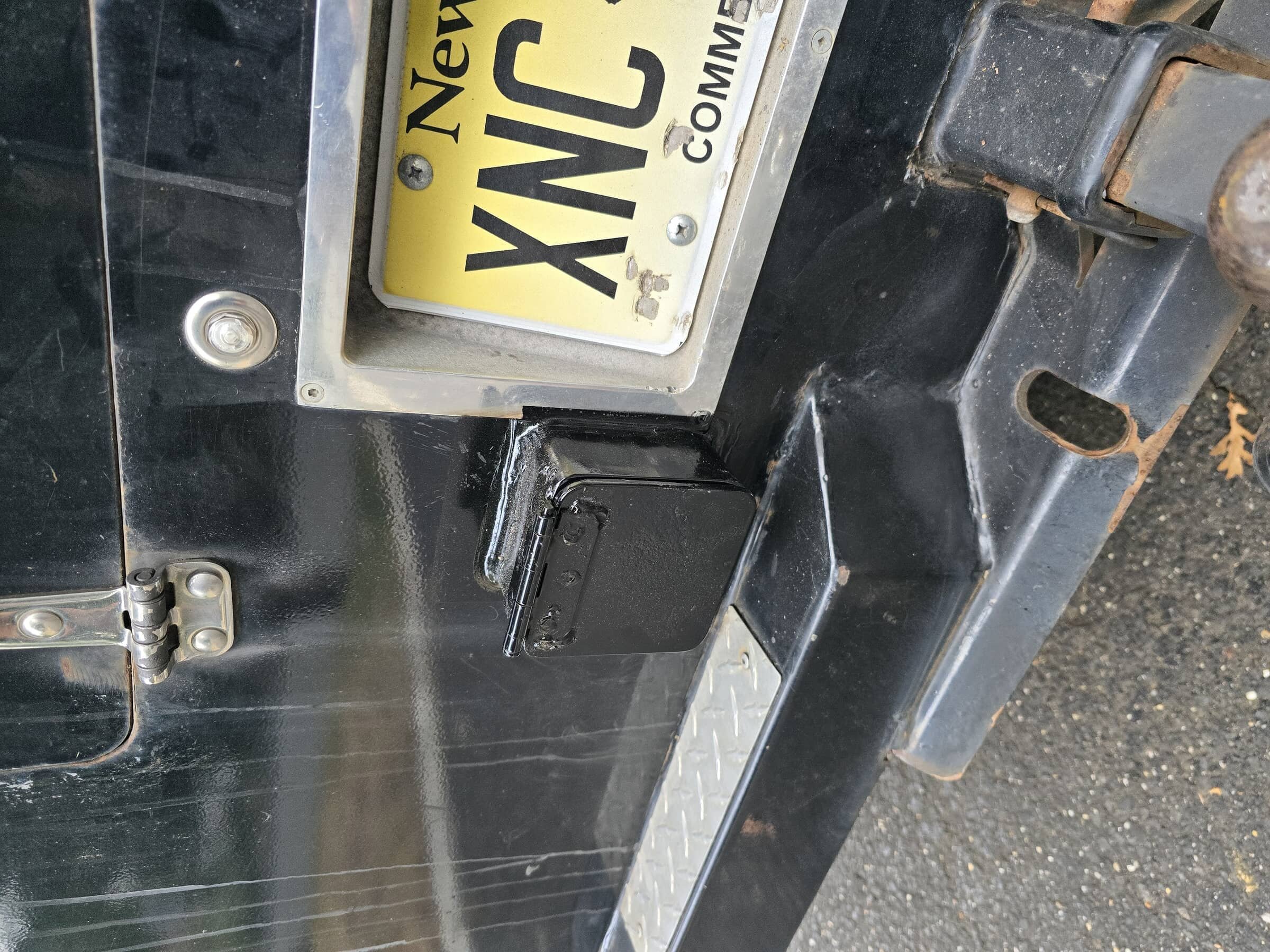 Close-up of a black metal vehicle bumper with a yellow license plate reading 'XNVNC,' a small black hinge, and a rusty hole in the metal.