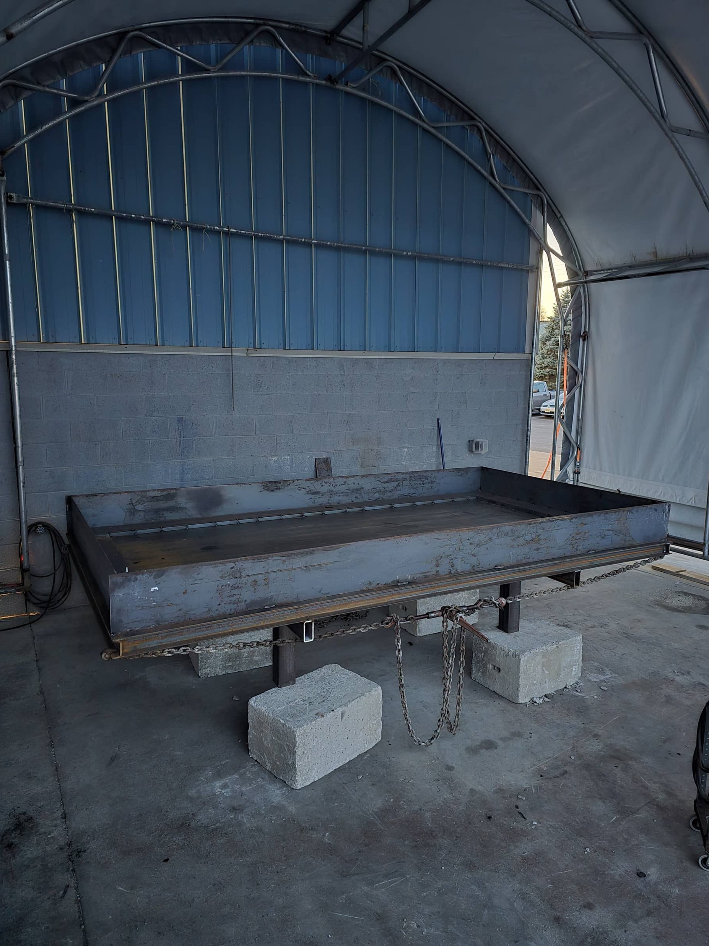 Large, rectangular metal grinding or polishing station with chains and concrete blocks for support, situated inside a spacious industrial tent with metal framing and concrete floor.
