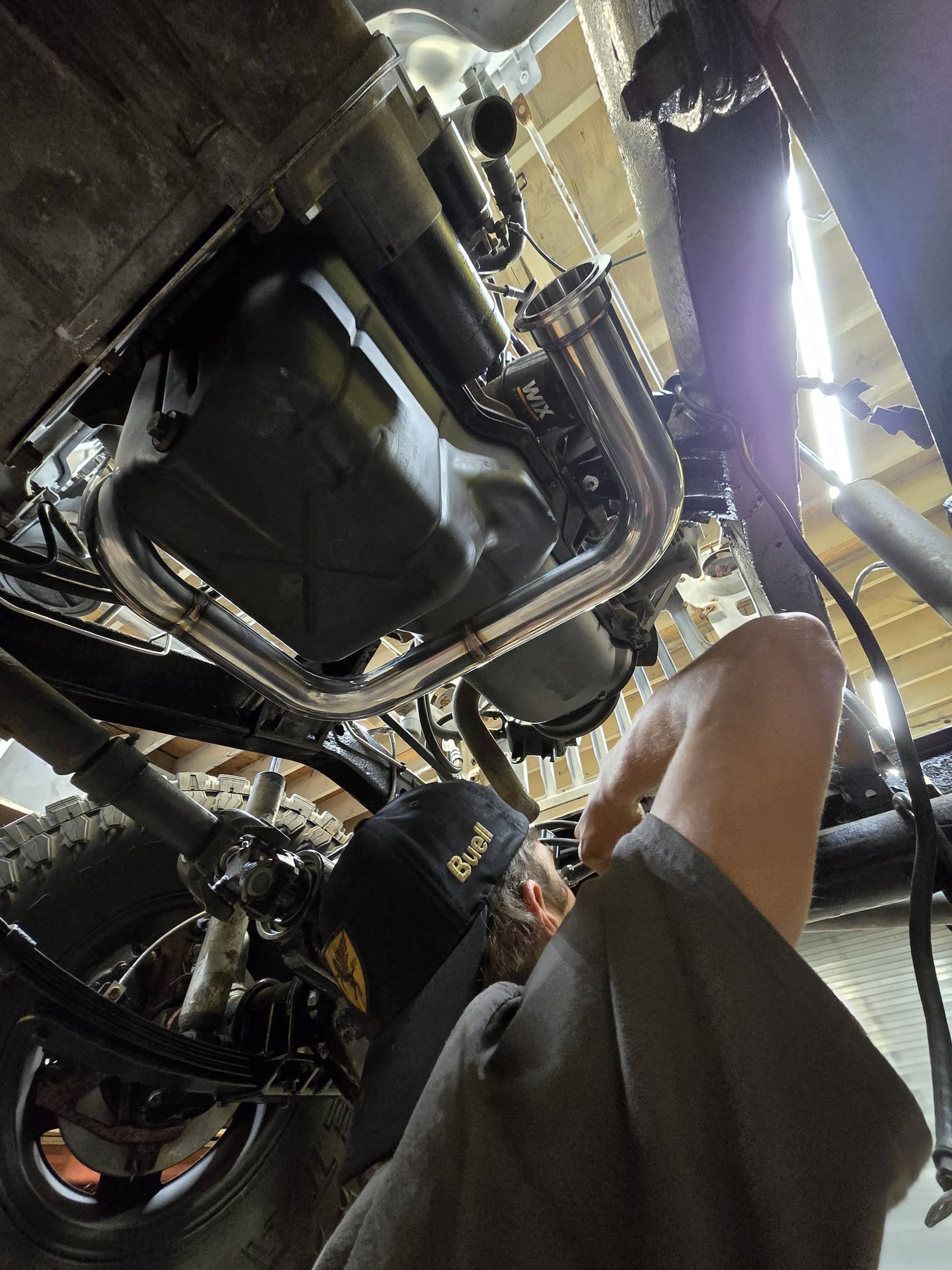 A person working underneath a vehicle, visible from the waist up, wearing a black cap and a gray shirt, performing maintenance or repair on the engine or chassis.