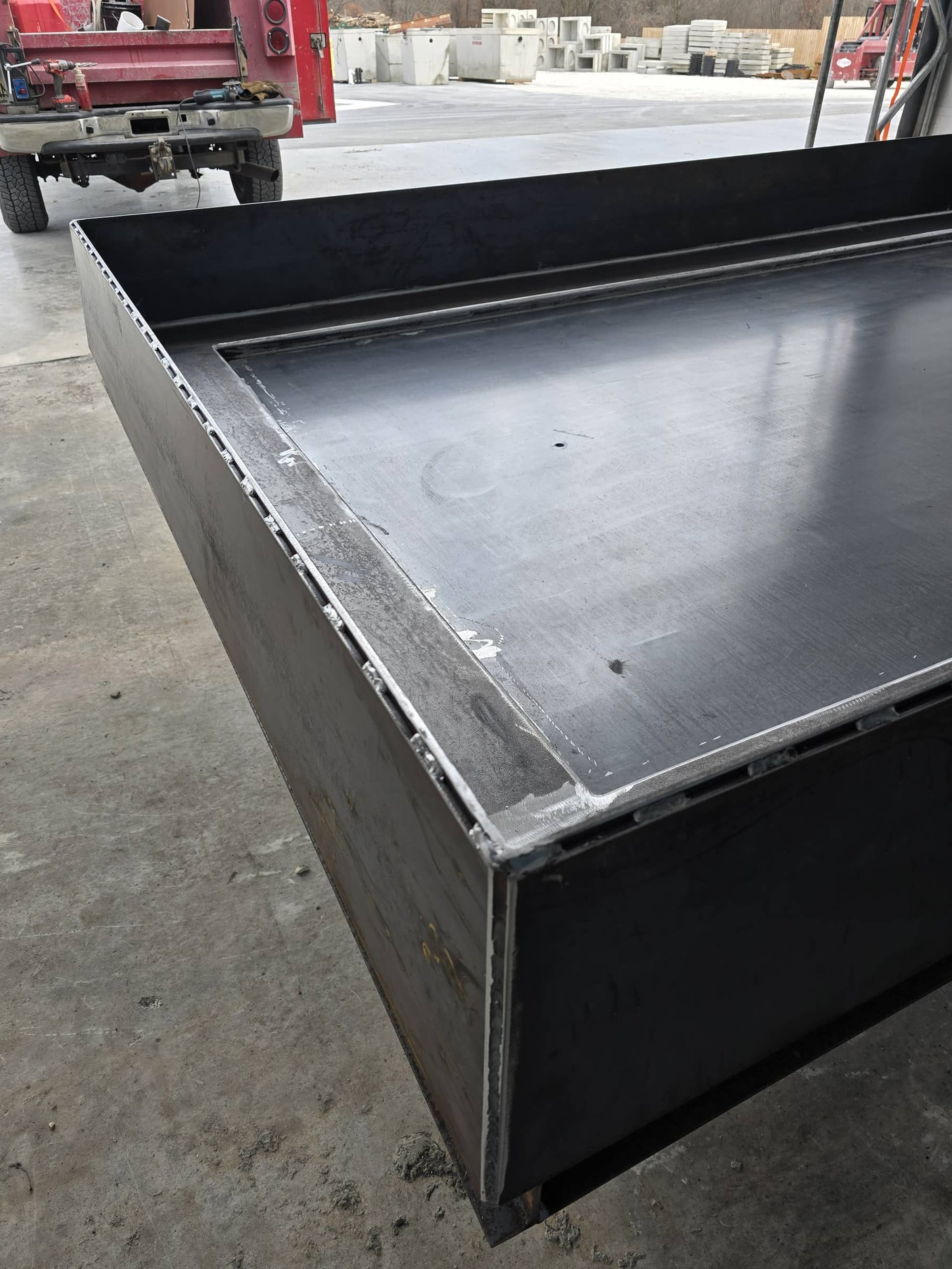 Close-up of a stainless steel industrial worktable or container with a smooth, reflective surface and a raised edge, situated in a workshop or construction site with concrete paving and construction materials in the background.
