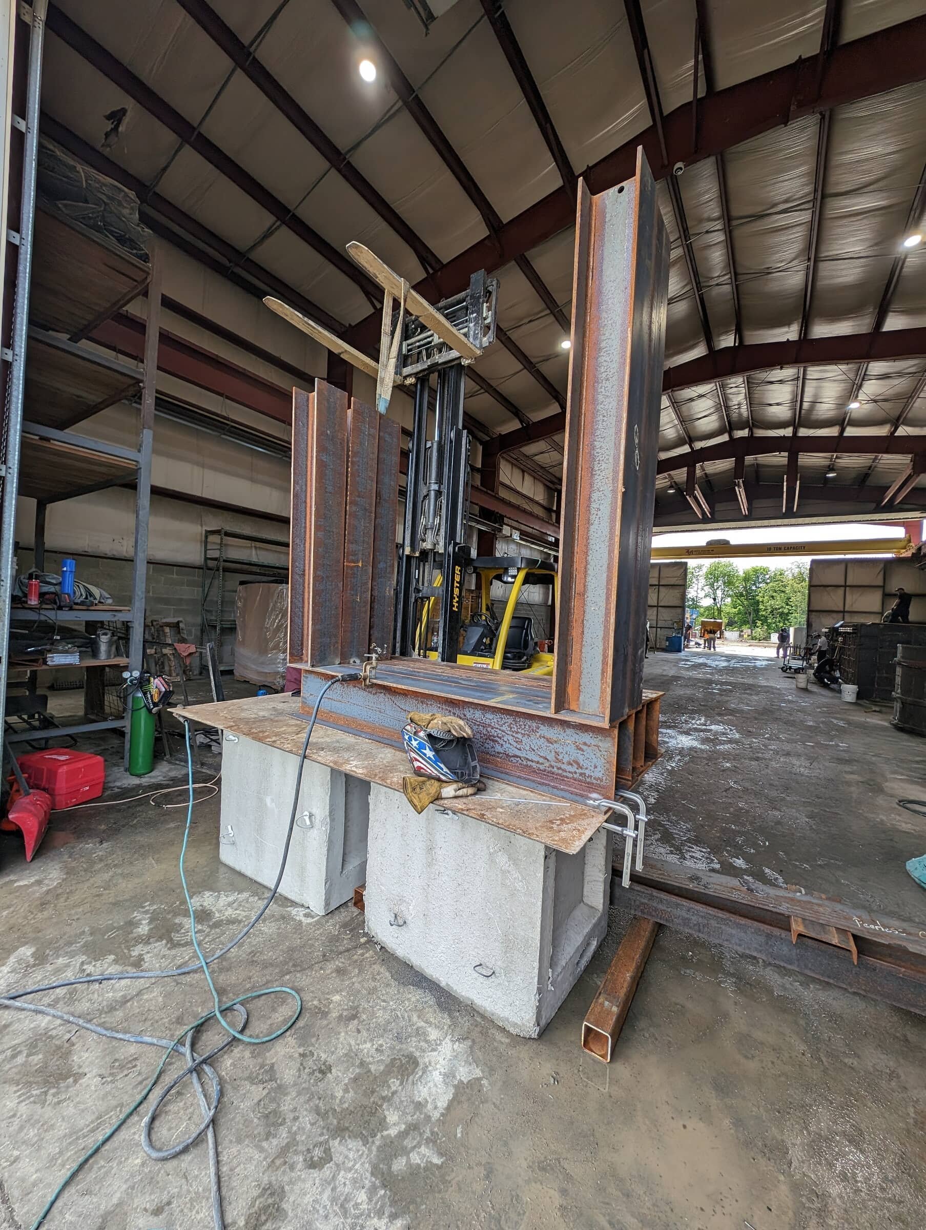 Large industrial steel frame structure being assembled inside a warehouse, supported by concrete blocks, with a forklift in the background.