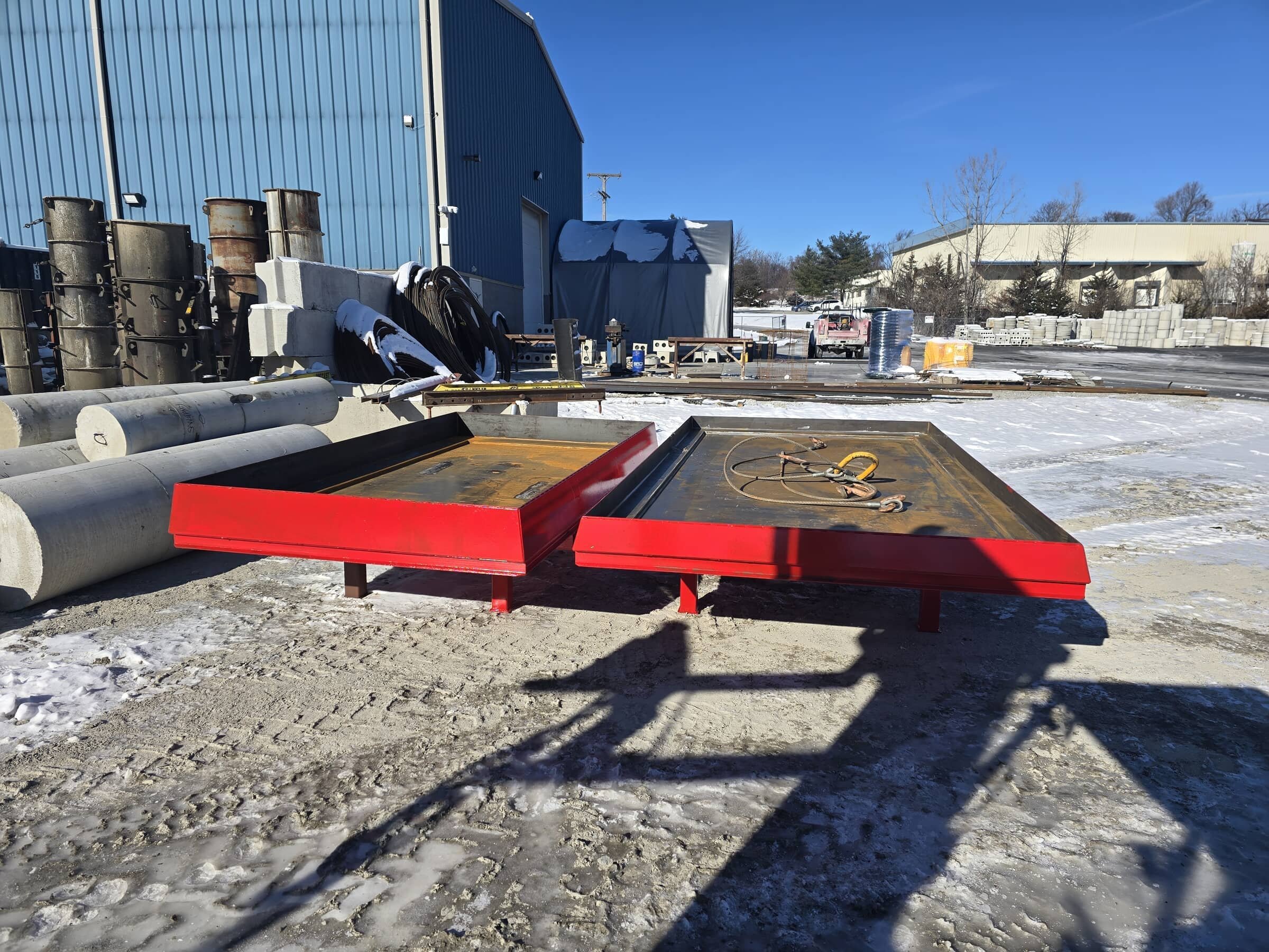 Industrial outdoor scene with large blue warehouse, concrete pipes, black hoses, metal cylinders, and two red platforms on snowy ground under clear blue sky.