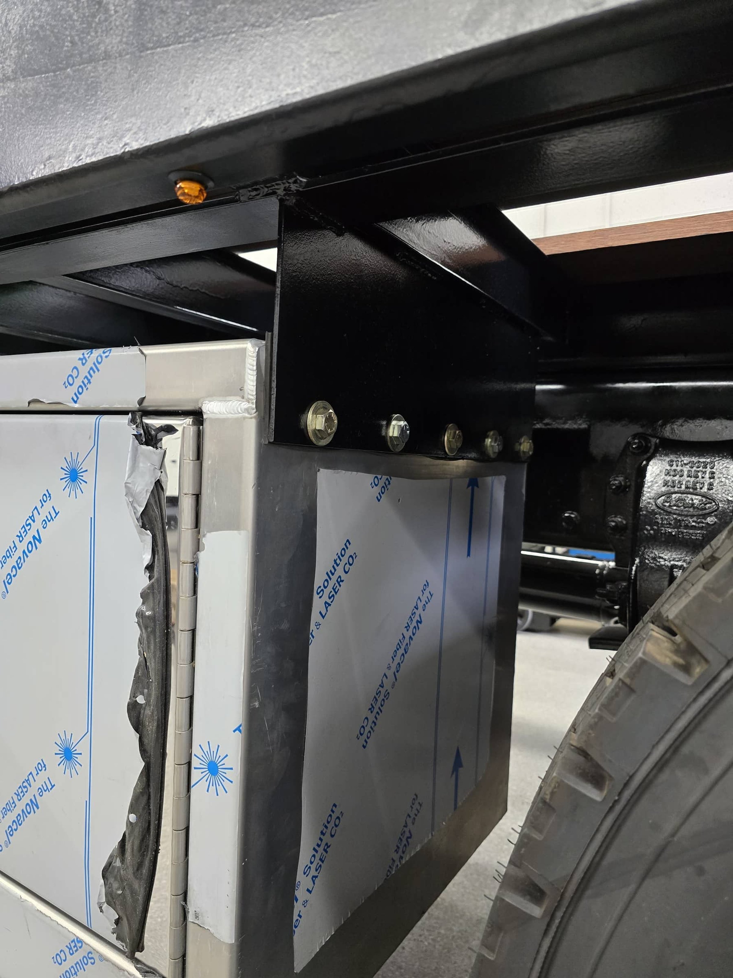 Close-up of a black metal support beam under a vehicle with a partially covered metal box attached to it, showing bolts securing the support and a tire partially visible on the right.