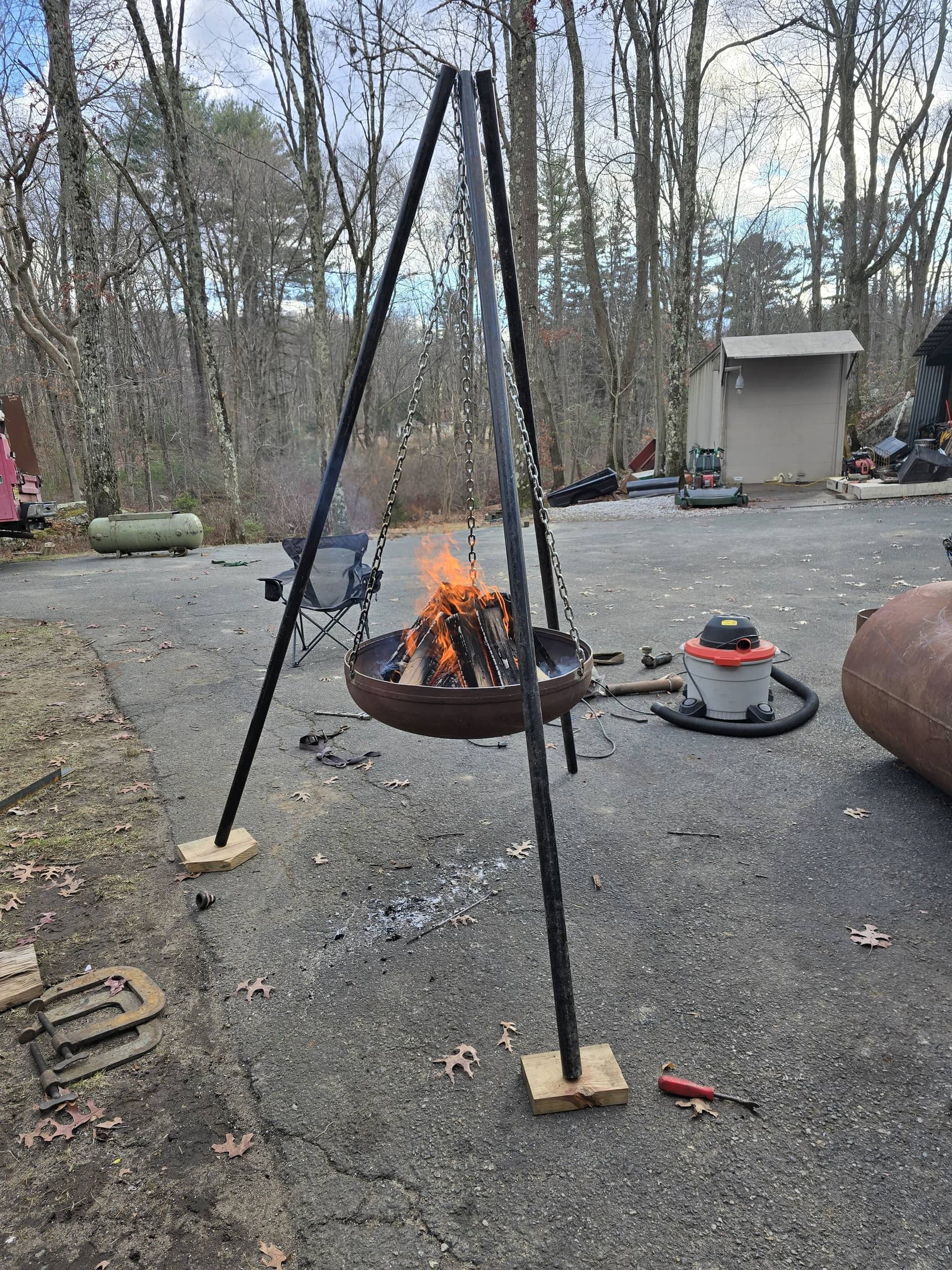 Outdoor scene with a hanging fire pit on a tripod stand burning wood, surrounded by tools, a vacuum, and a backyard with trees and storage shed.