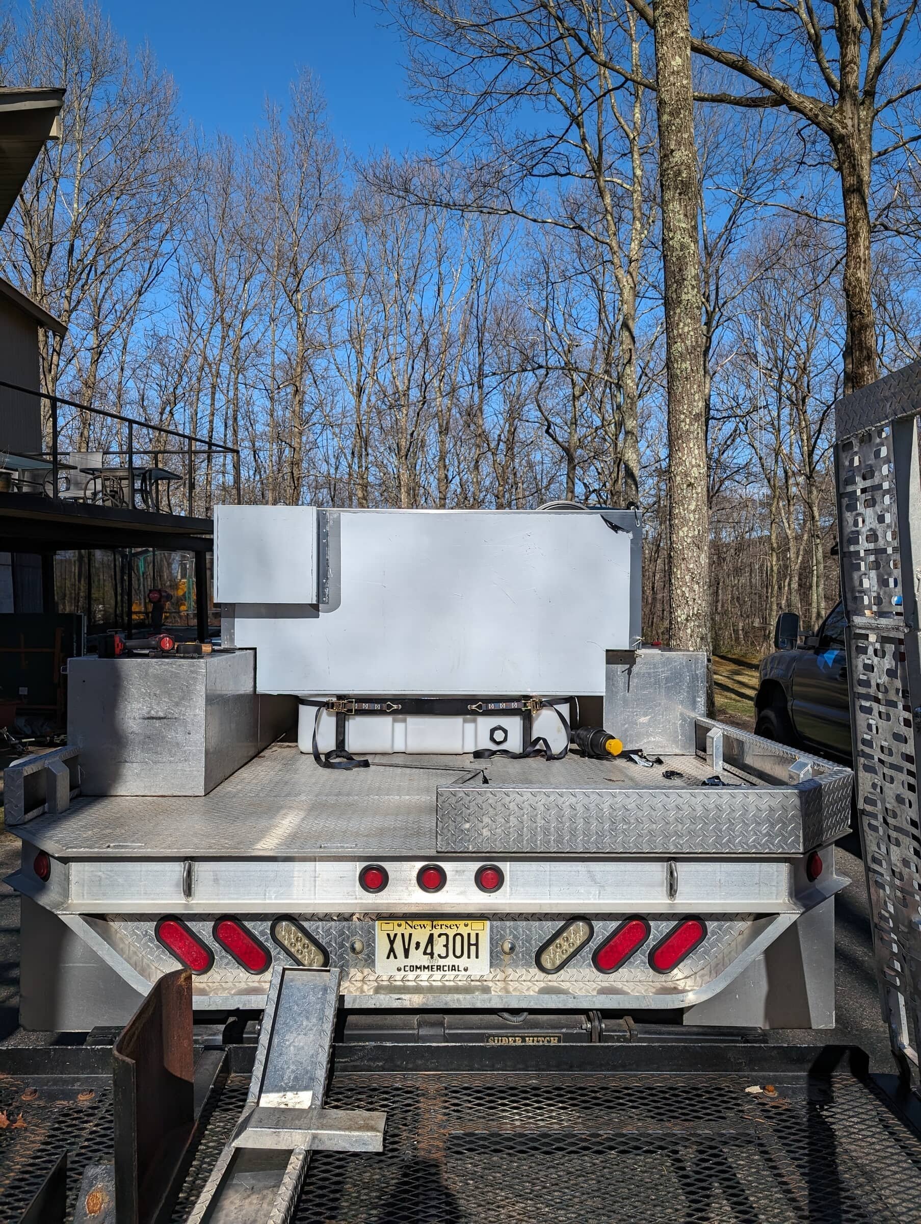 Rear view of a utility truck with a metal flatbed, equipped with storage boxes, straps, and tools, parked outdoors with trees and a building visible in the background.