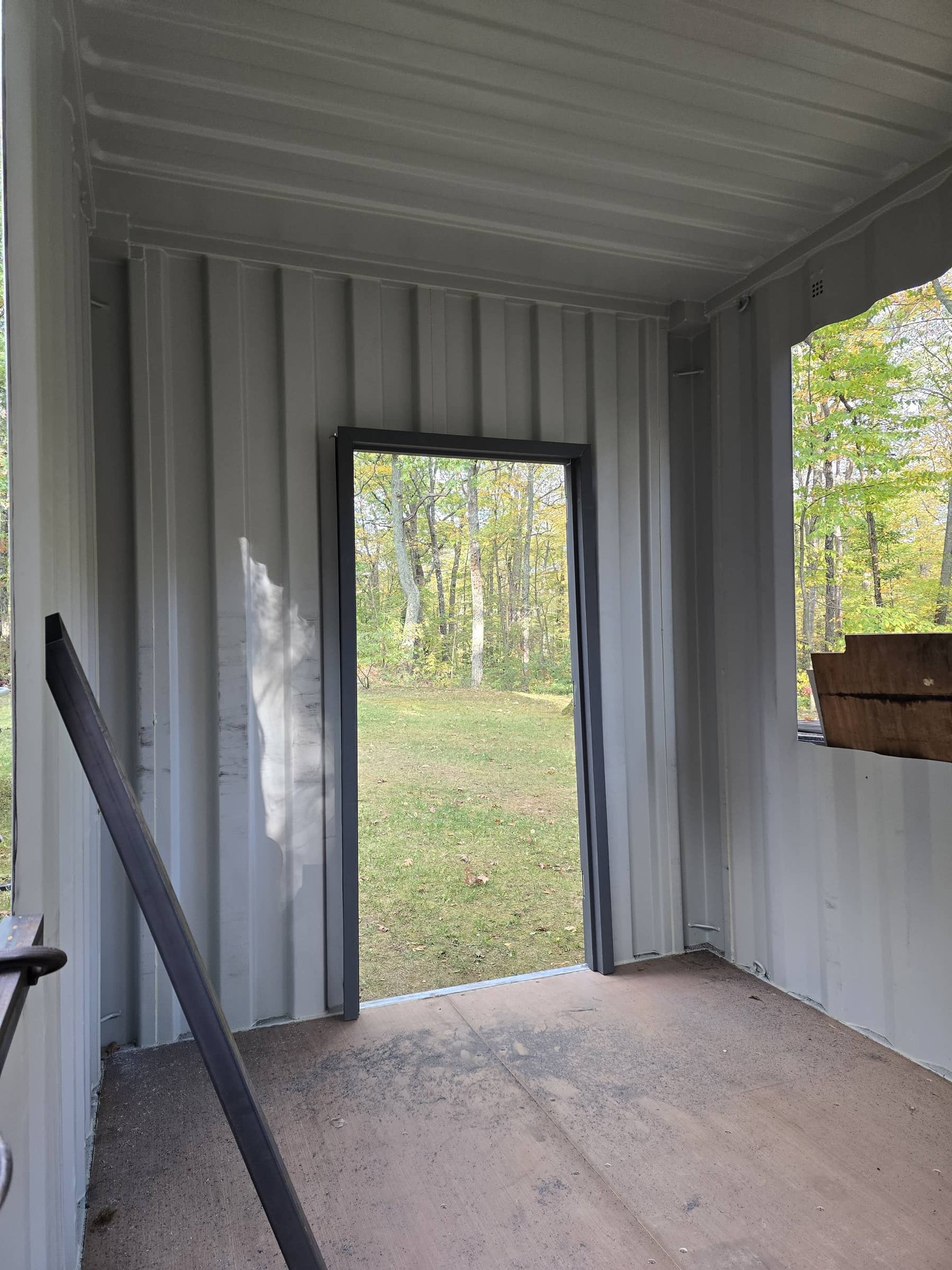 Interior of a small metal shed with a partially open door revealing a grassy yard and trees outside.