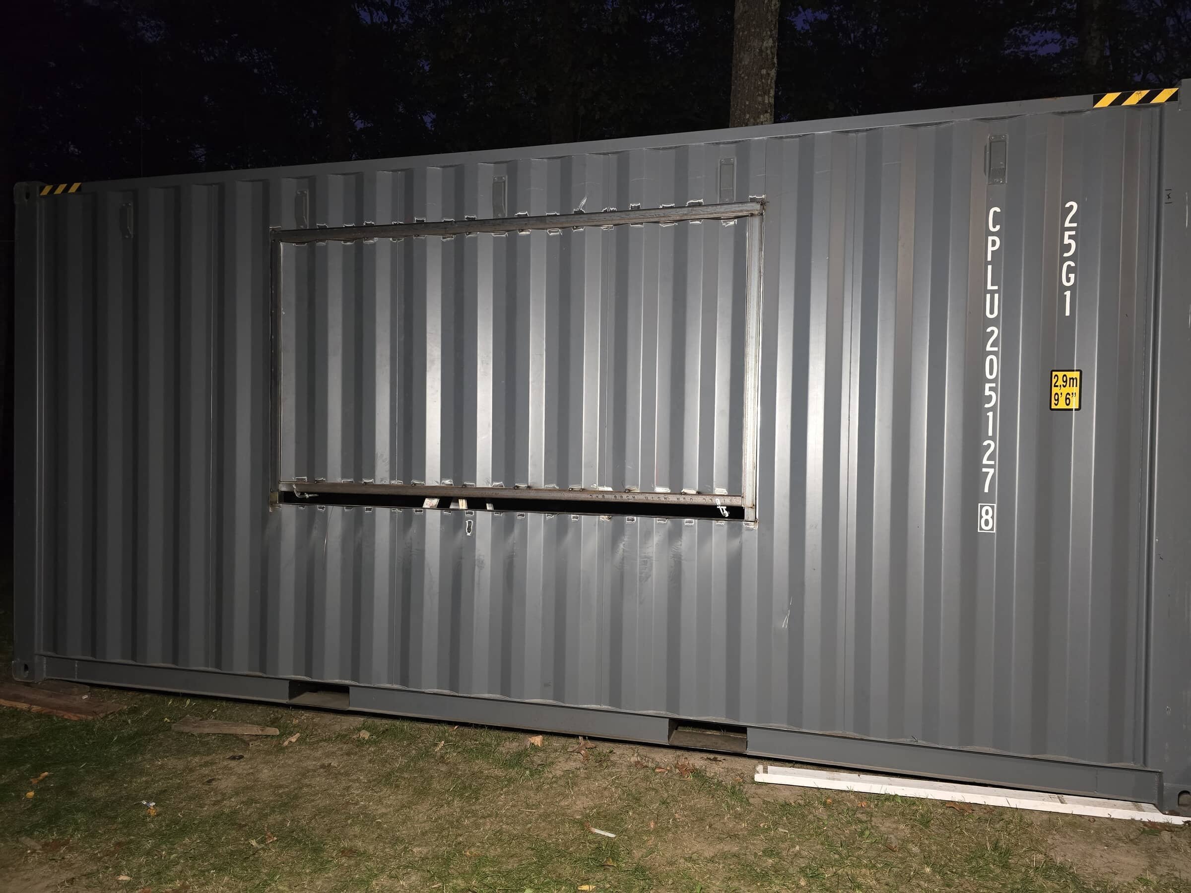 A large gray shipping container with a missing side panel, revealing an open window frame, placed on grass. The container has white and yellow markings with barcode and measurement labels.