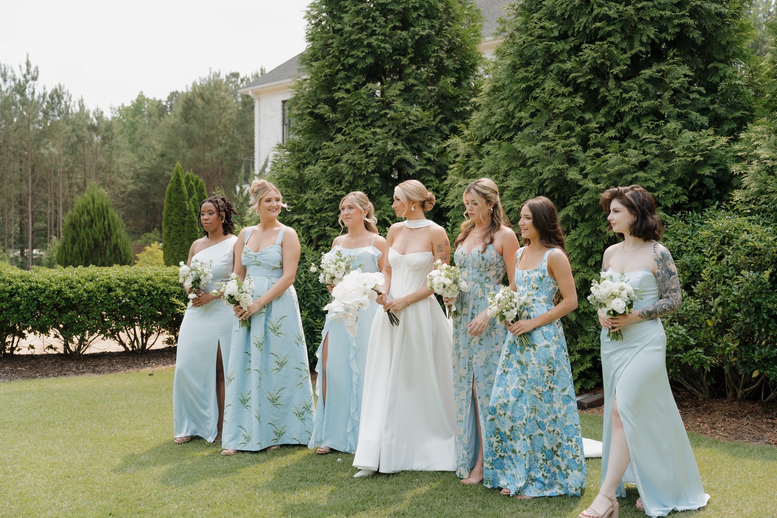 A bride and seven bridesmaids standing outdoors on a lawn with trees and a house in the background, holding bouquets of white flowers, dressed in white and light blue dresses.