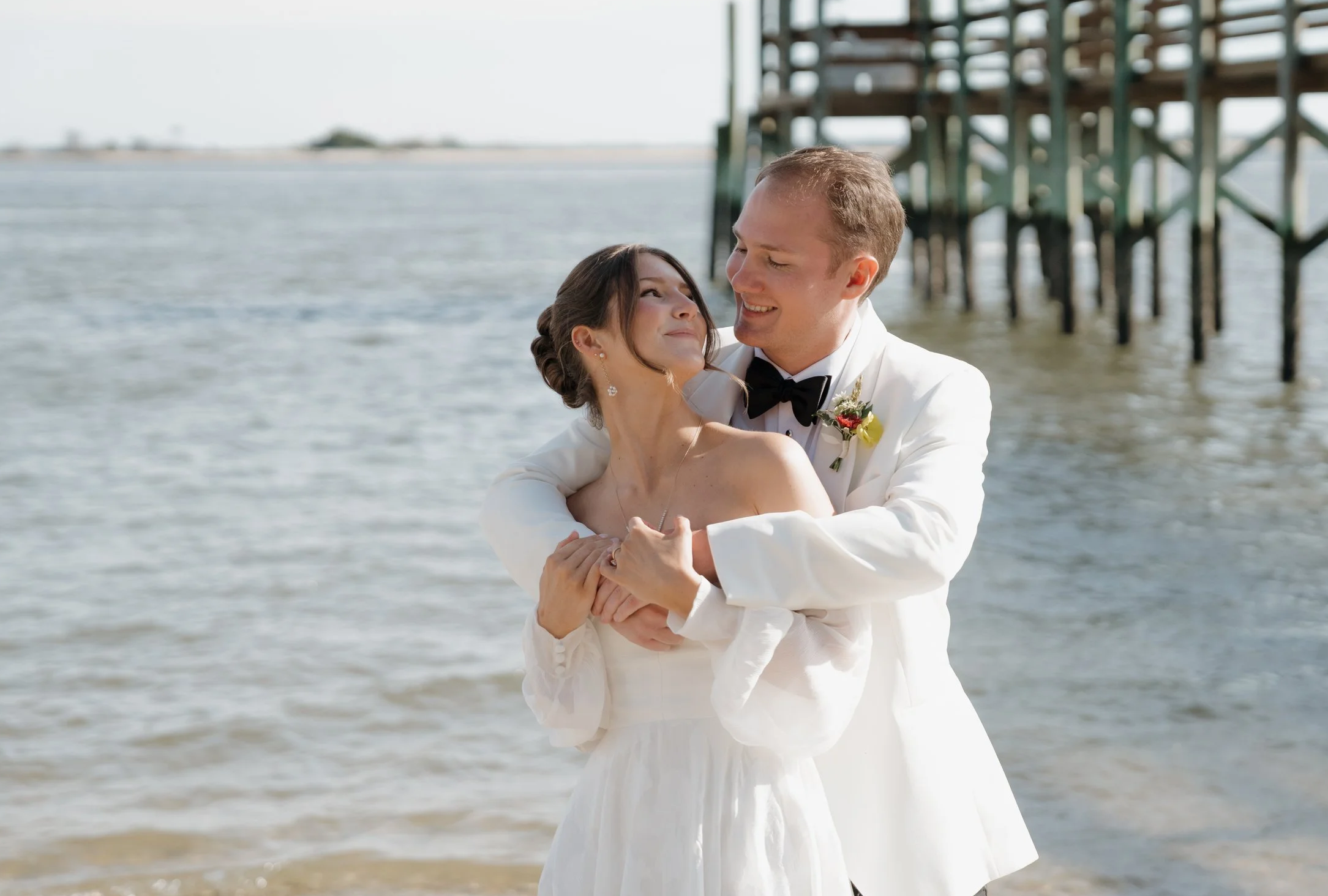 A bride and groom happily embracing on a beach, with the groom in a white tuxedo and the bride in a white wedding dress, standing near a pier.