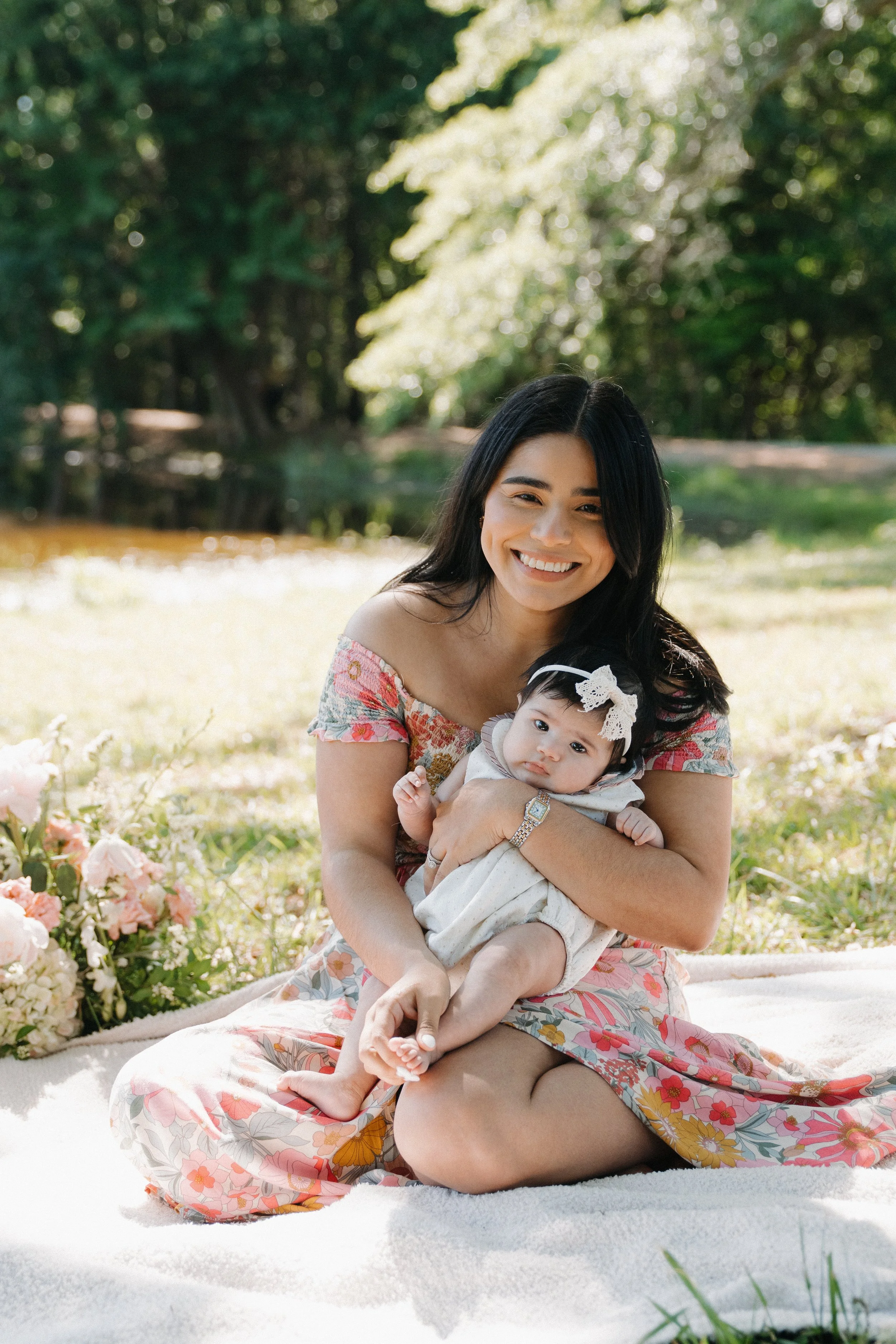 A woman and a baby sitting on a blanket outdoors in a park, surrounded by flowers and greenery, smiling at the camera.