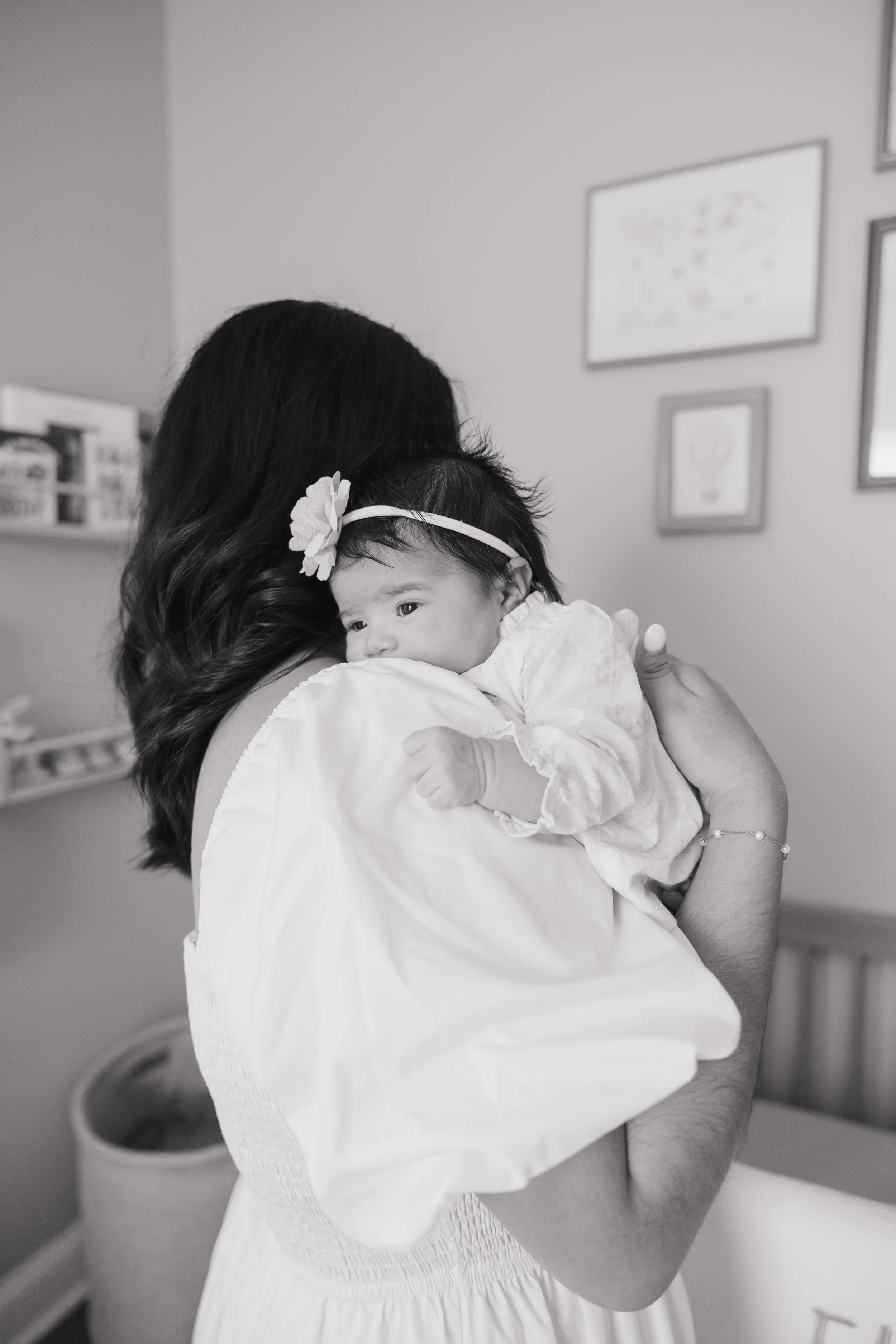 A woman holding a baby in a home setting. The woman has long, wavy hair and is wearing a light-colored outfit. The baby has a headband with a flower and is resting against the woman's shoulder, looking at the camera. The background shows a shelf and framed pictures on the wall.