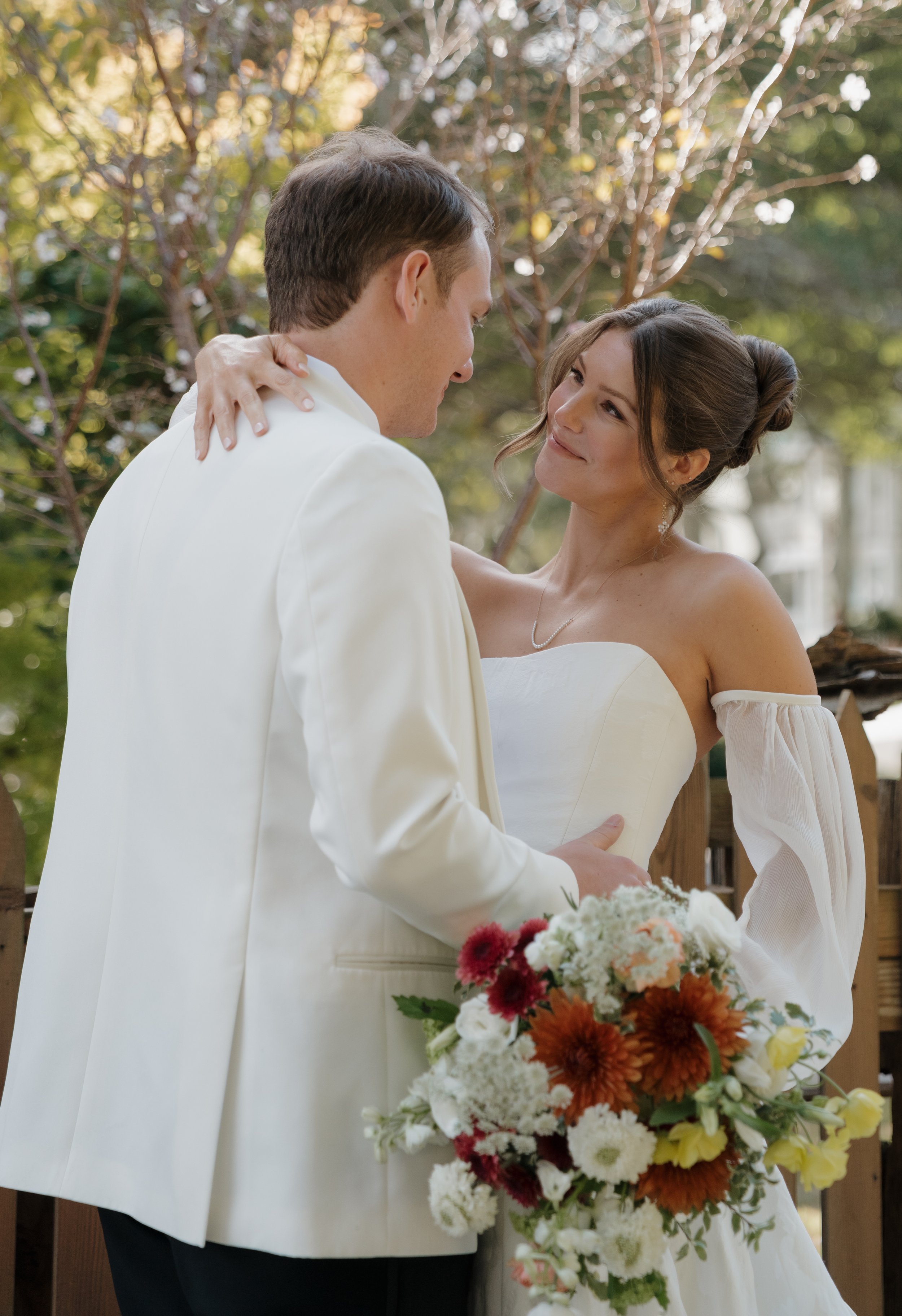 A bride and groom sharing a loving moment outdoors, holding each other, with the bride holding a bouquet of colorful flowers.