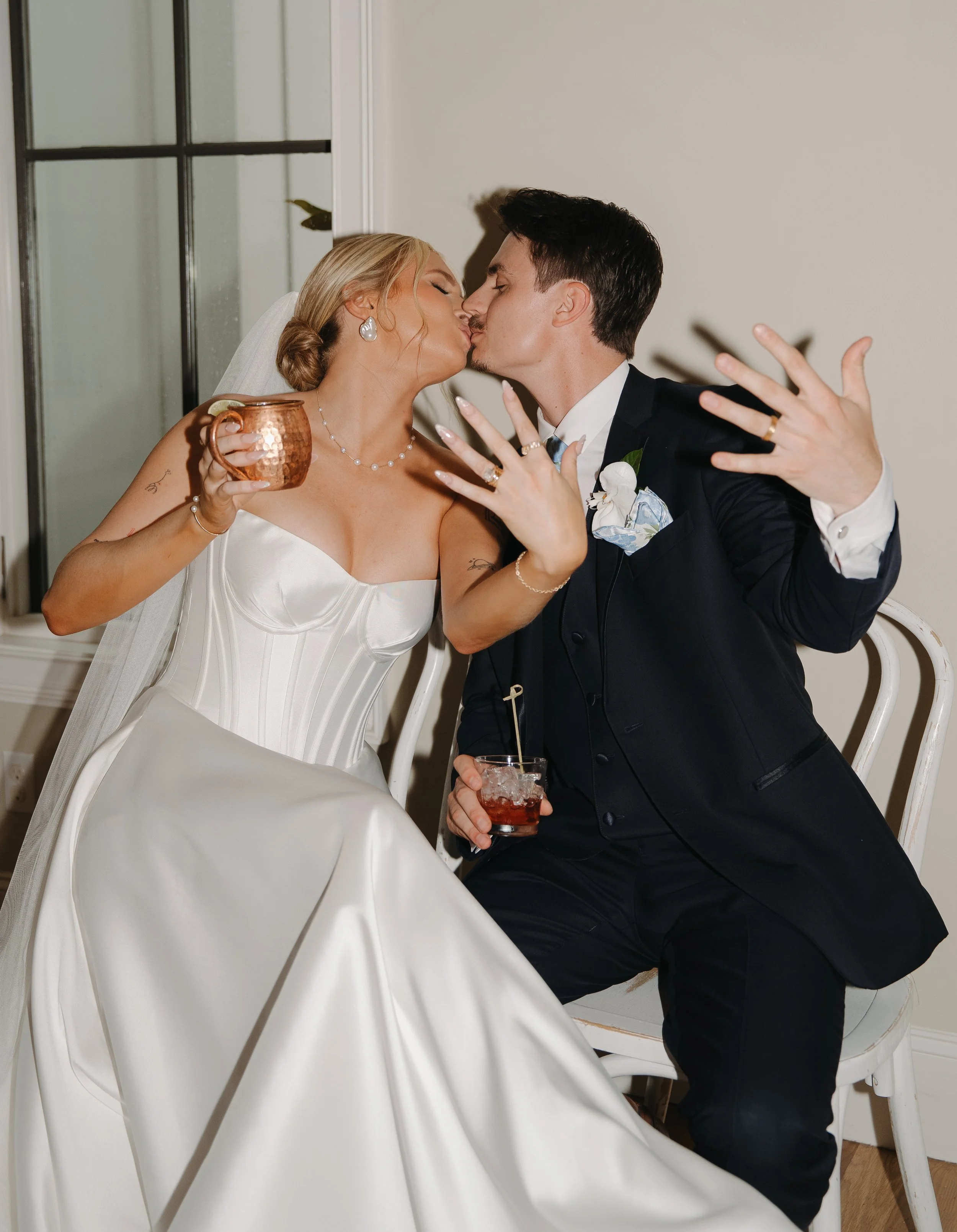 Bride and groom kissing at their wedding, with drinks in hand, sitting on chairs indoors.
Reception wedding photo.