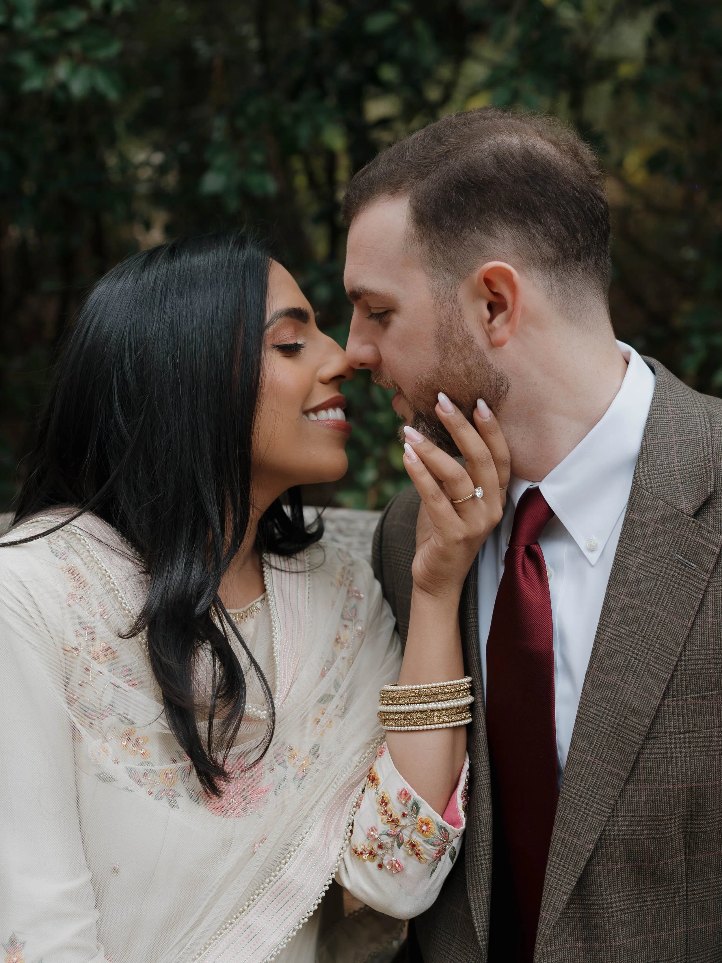 A couple shares an intimate moment, about to touch noses, outdoors with greenery in the background.