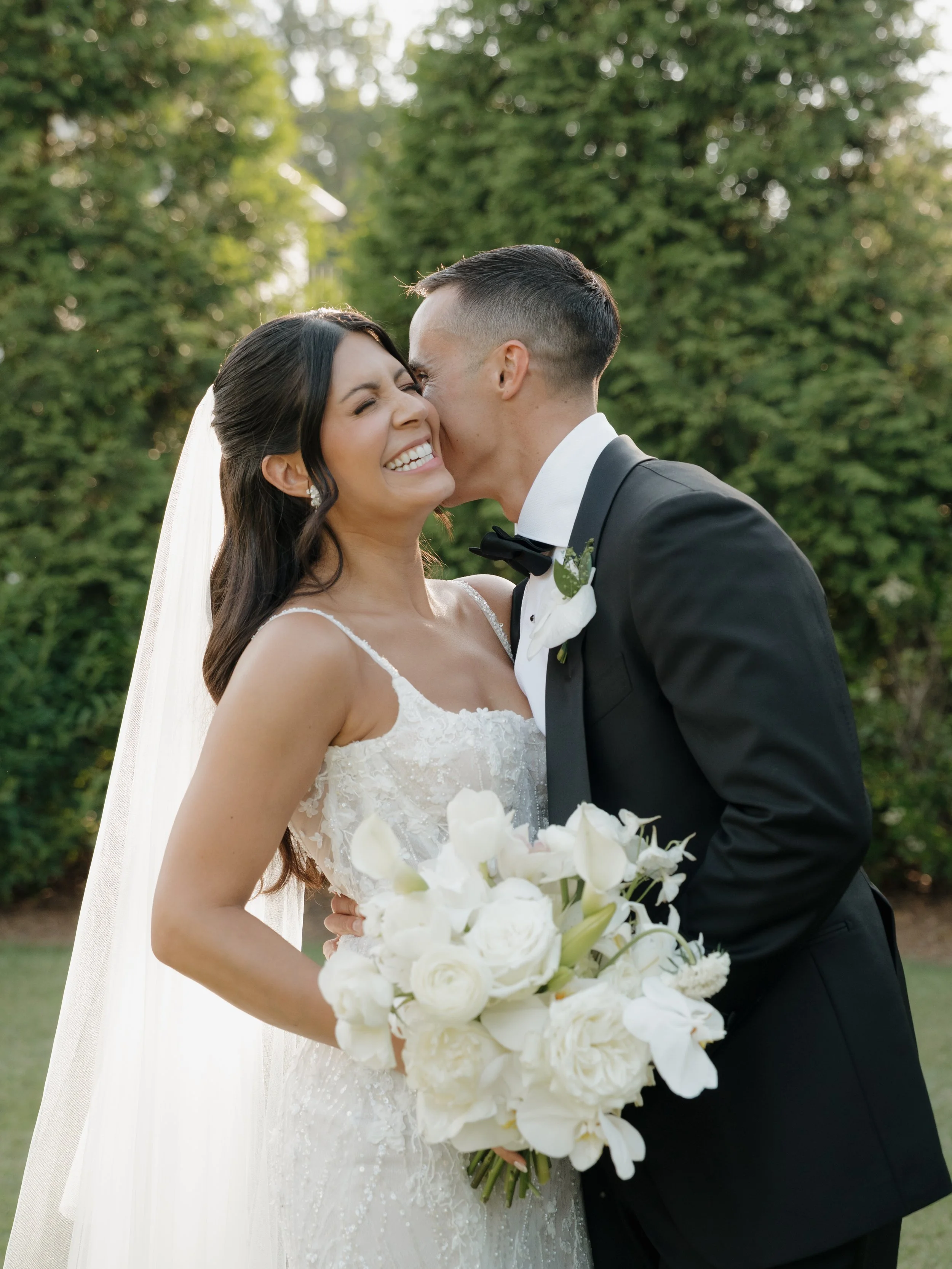 A bride and groom sit close together in a vintage car, holding hands with their eyes closed; she wears a wedding dress and veil, he in a suit.