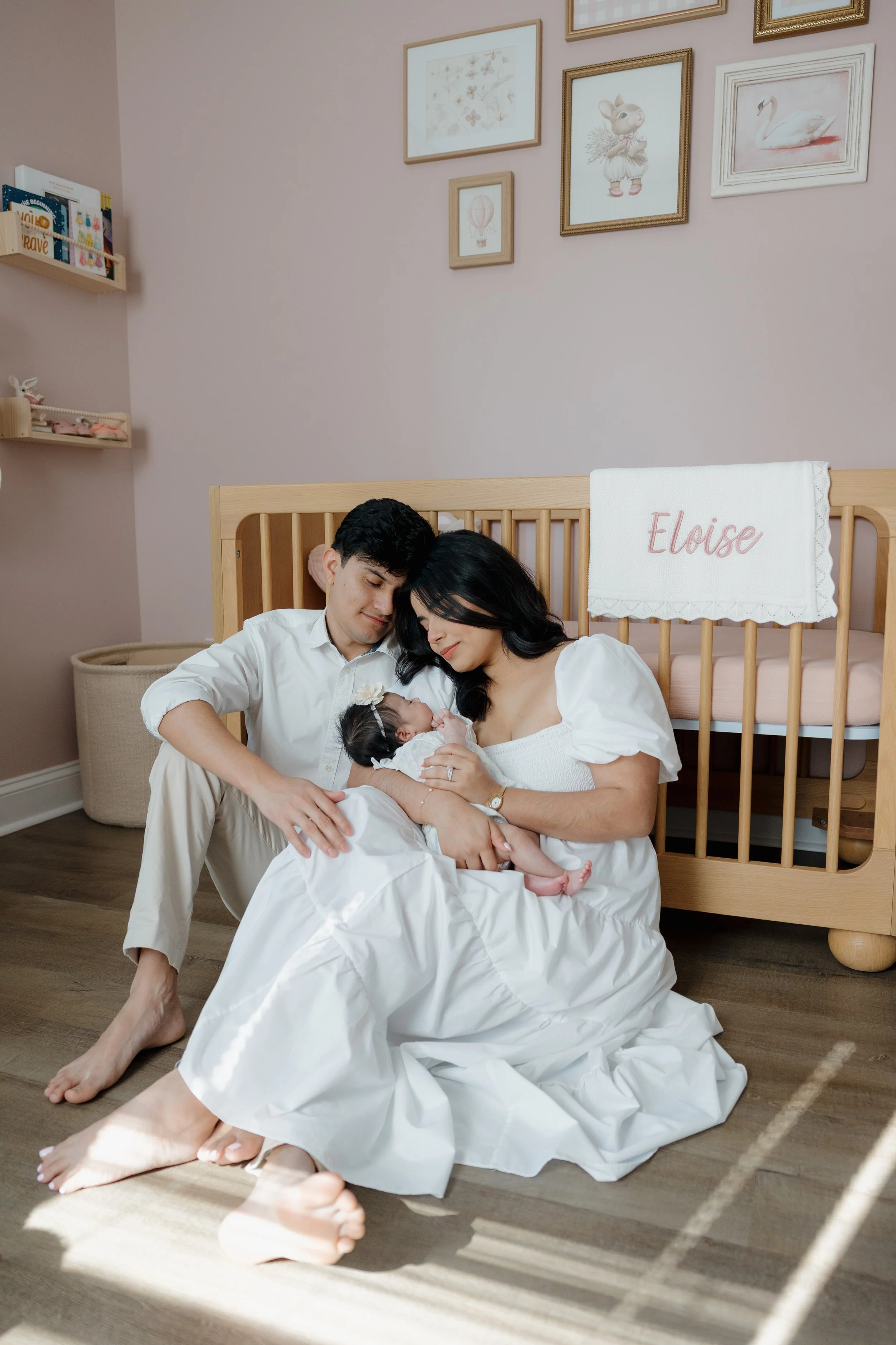 A couple dressed in white robes sitting on the floor, holding a newborn baby, smiling and looking at the baby. Behind them, a wooden crib with a personalized name tag 'Eloise' is visible. The room has light pink walls decorated with framed artwork including a mouse illustration, a swan, and other images. Soft natural light illuminates the scene.