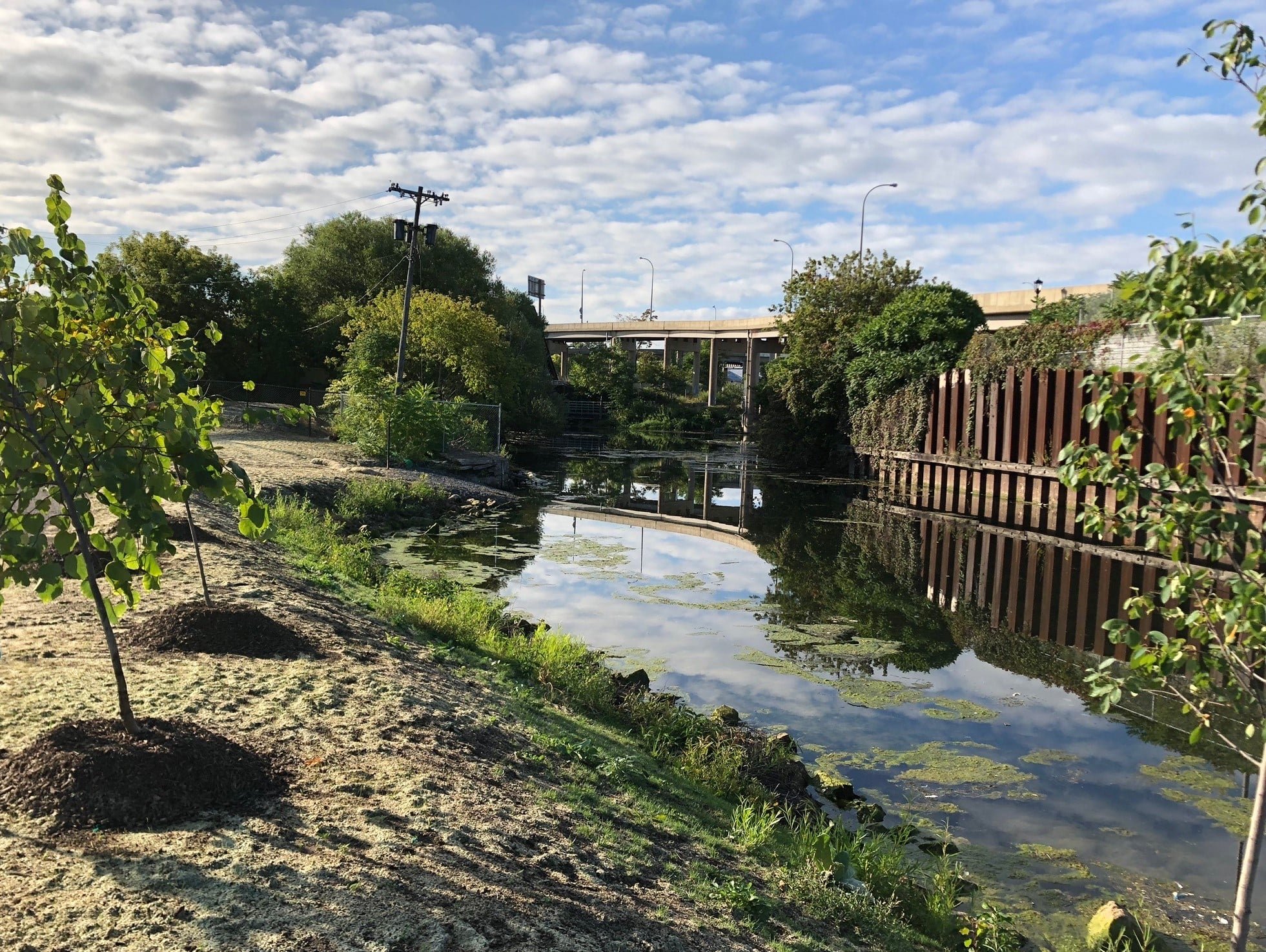 A small river or canal with a grassy bank, lined with young trees and bushes. The water reflects the sky and surrounding greenery. In the background, there's an overpass bridge and utility poles under a partly cloudy sky.
