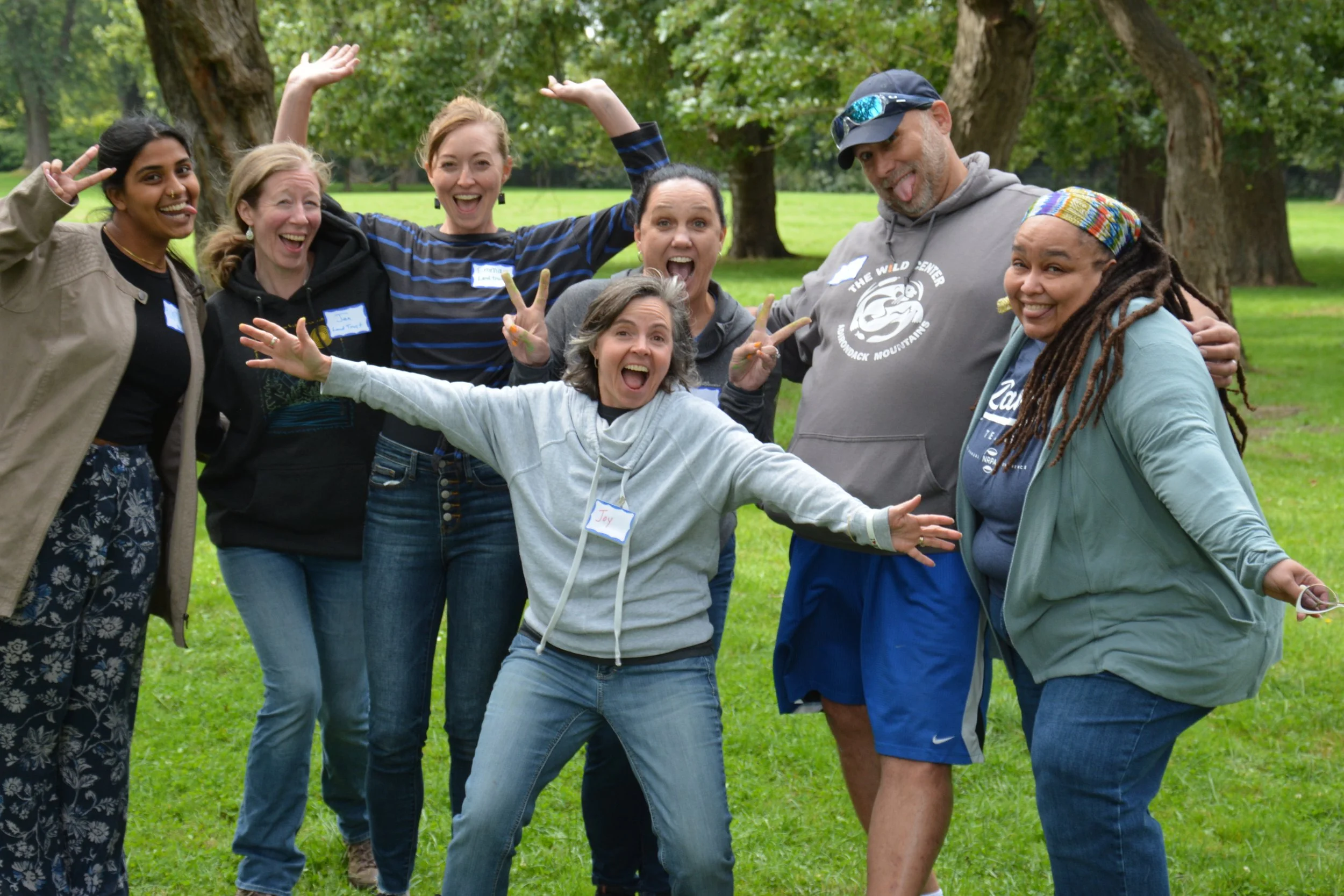 Group of seven people posing playfully outdoors, all smiling, set in a green grassy area with trees in the background.