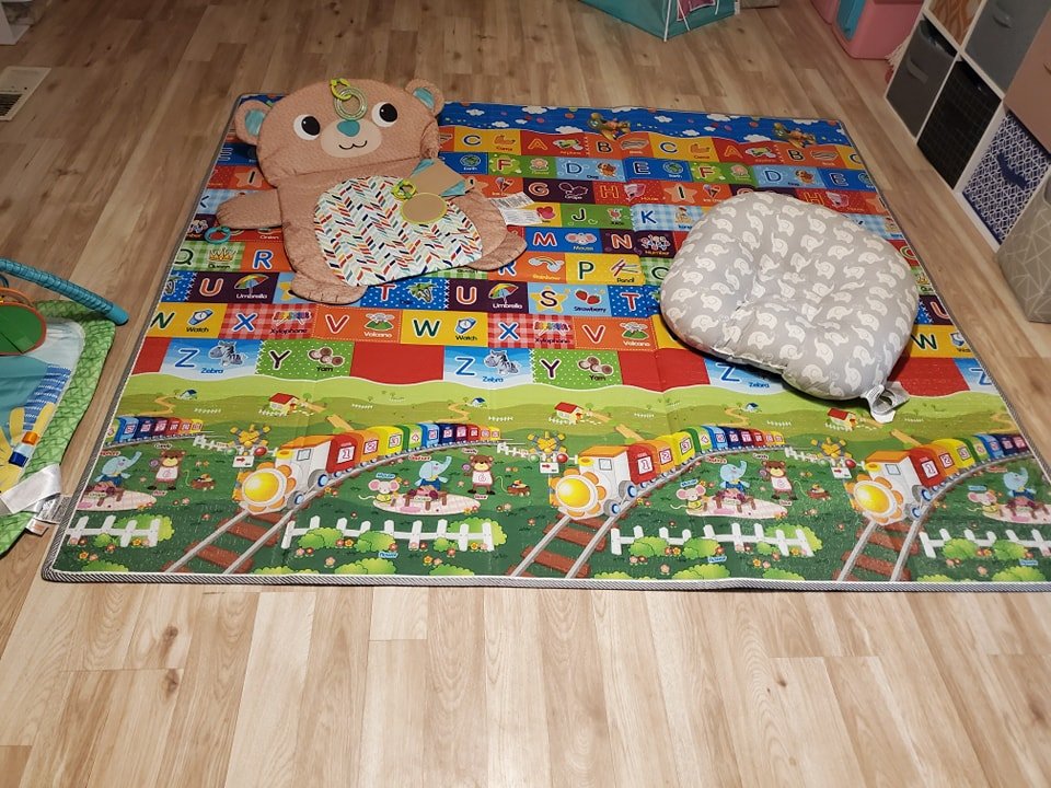 Colorful children's playmat with alphabet and train illustrations on a wooden floor, featuring a plush cushion and a large teddy bear design.