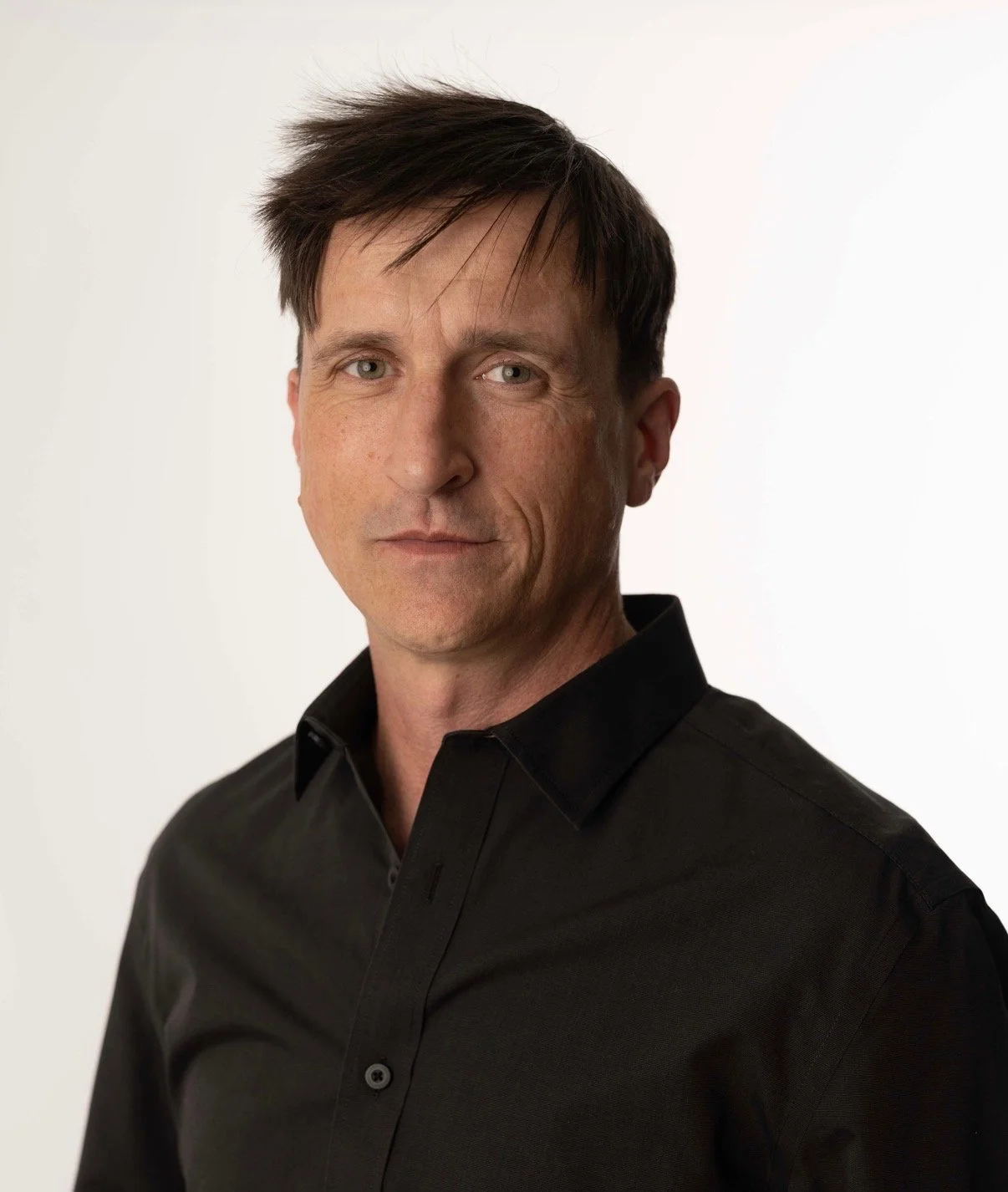 A man with short, dark hair and a slight beard, wearing a black shirt, standing outside in an urban setting with modern glass buildings and trees in the background.