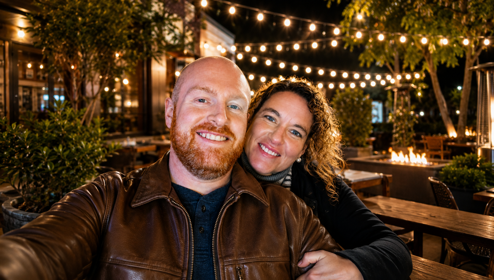 A smiling couple taking a selfie at an outdoor restaurant or patio at night, with string lights overhead and a fire pit in the background.