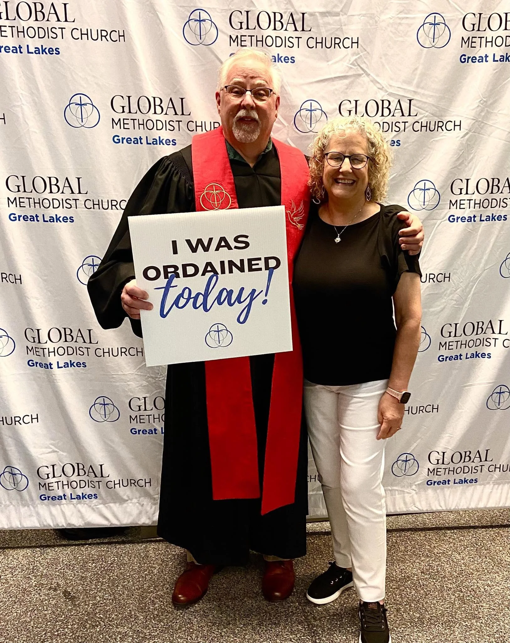 A man dressed in religious robes holding a sign that reads 'I WAS ORDAINED today!', standing next to a woman. They are in front of a backdrop with the logo and name of the Global Methodist Church and Great Lakes.