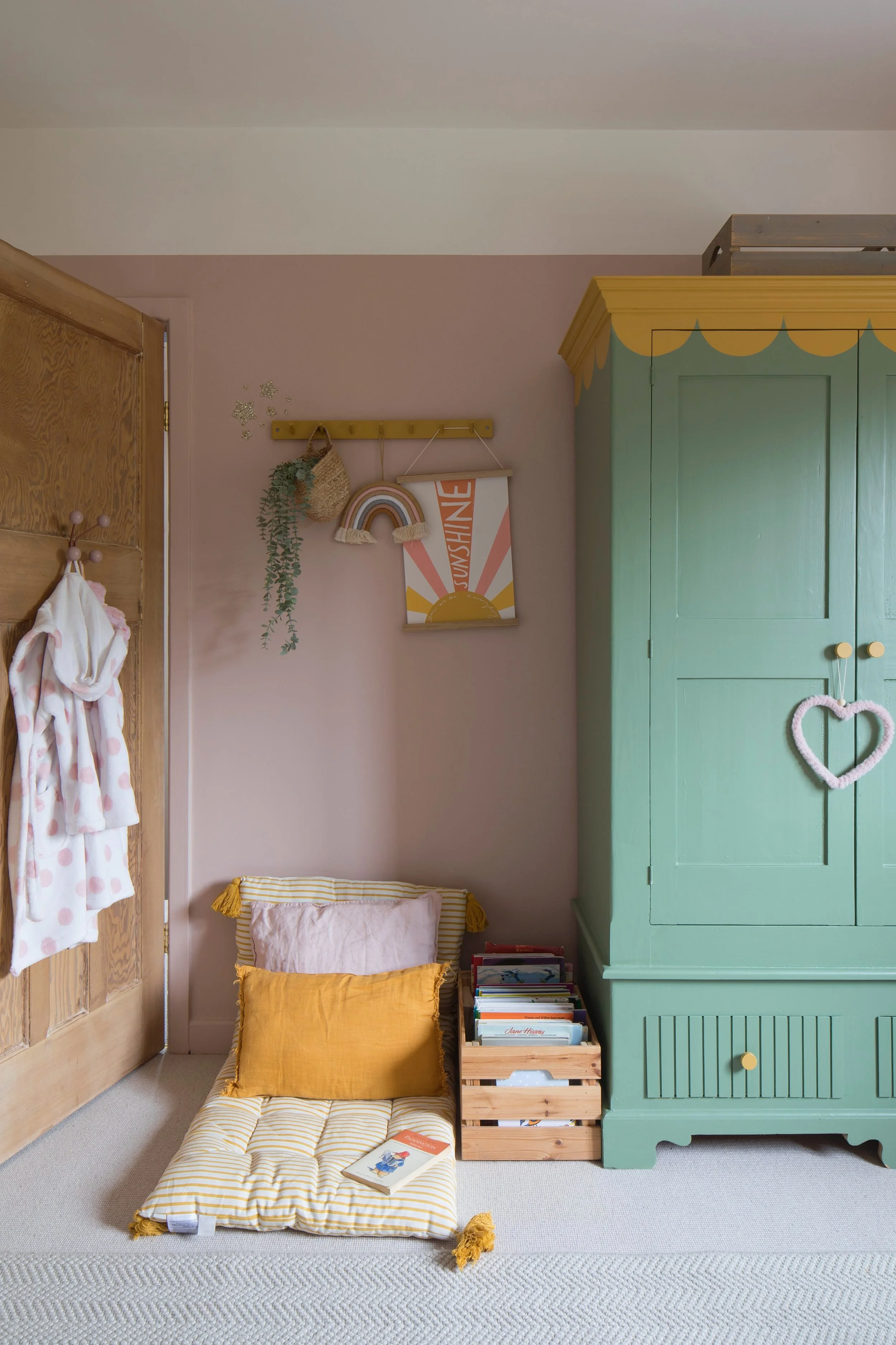 A cozy corner in a child's room with a green wardrobe decorated with a heart-shaped hanging. Next to it, a stack of wooden crates with books. A cushioned floor seat with striped yellow fabric and pillows is positioned against a pink wall. Above, a rack holds a woven basket, a rainbow decoration, and a colorful "SUNSHINE" poster. A pink robe with polka dots hangs on a door, partially visible on the left. The room has light-colored walls and beige carpet flooring.