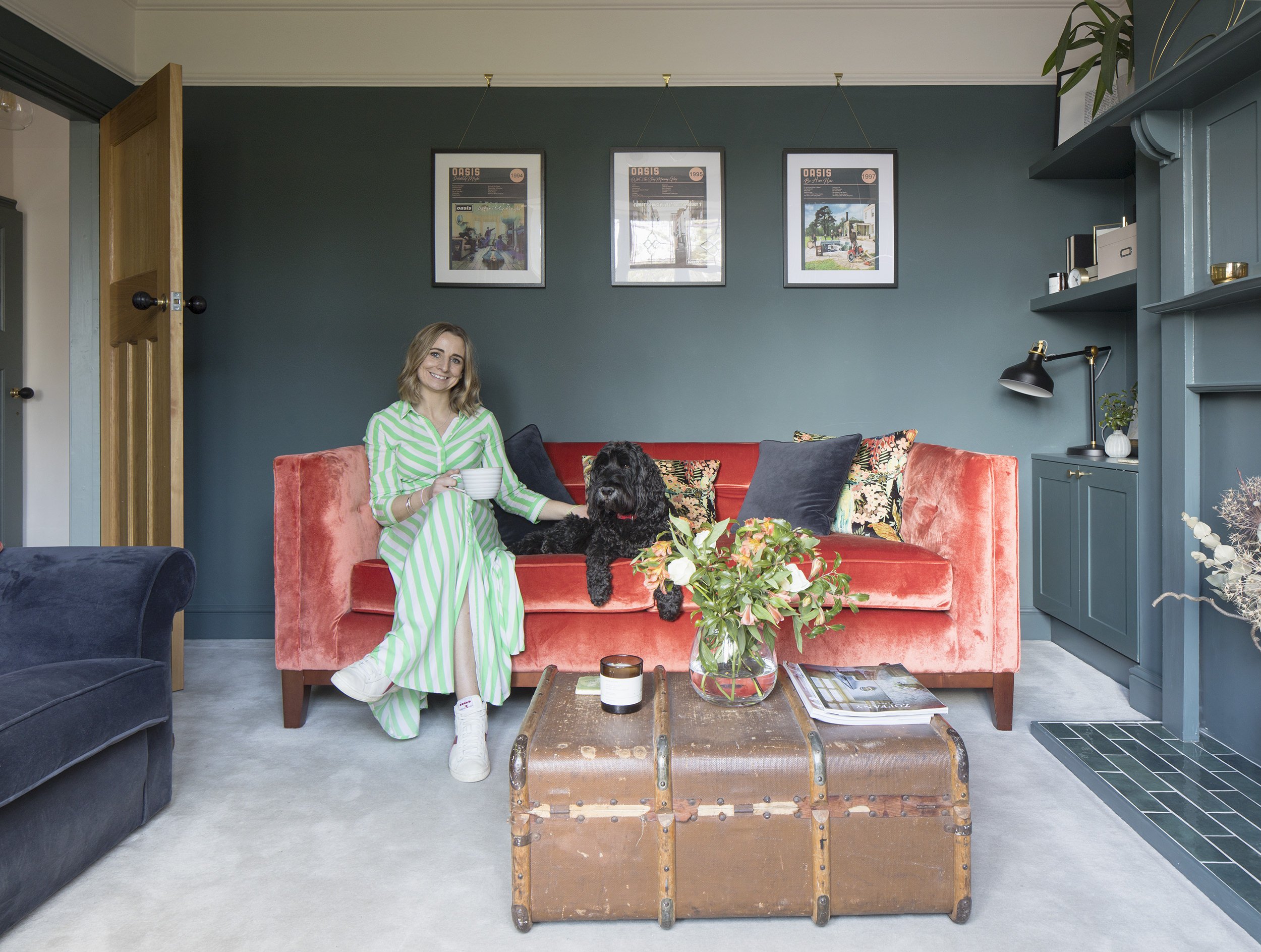 Erica Burton of Studio Dee on orange velvet sofa holding a mug, with a black dog next to her. The room has dark green walls decorated with framed pictures. There's a wooden chest as a coffee table with flowers and magazines on top.
