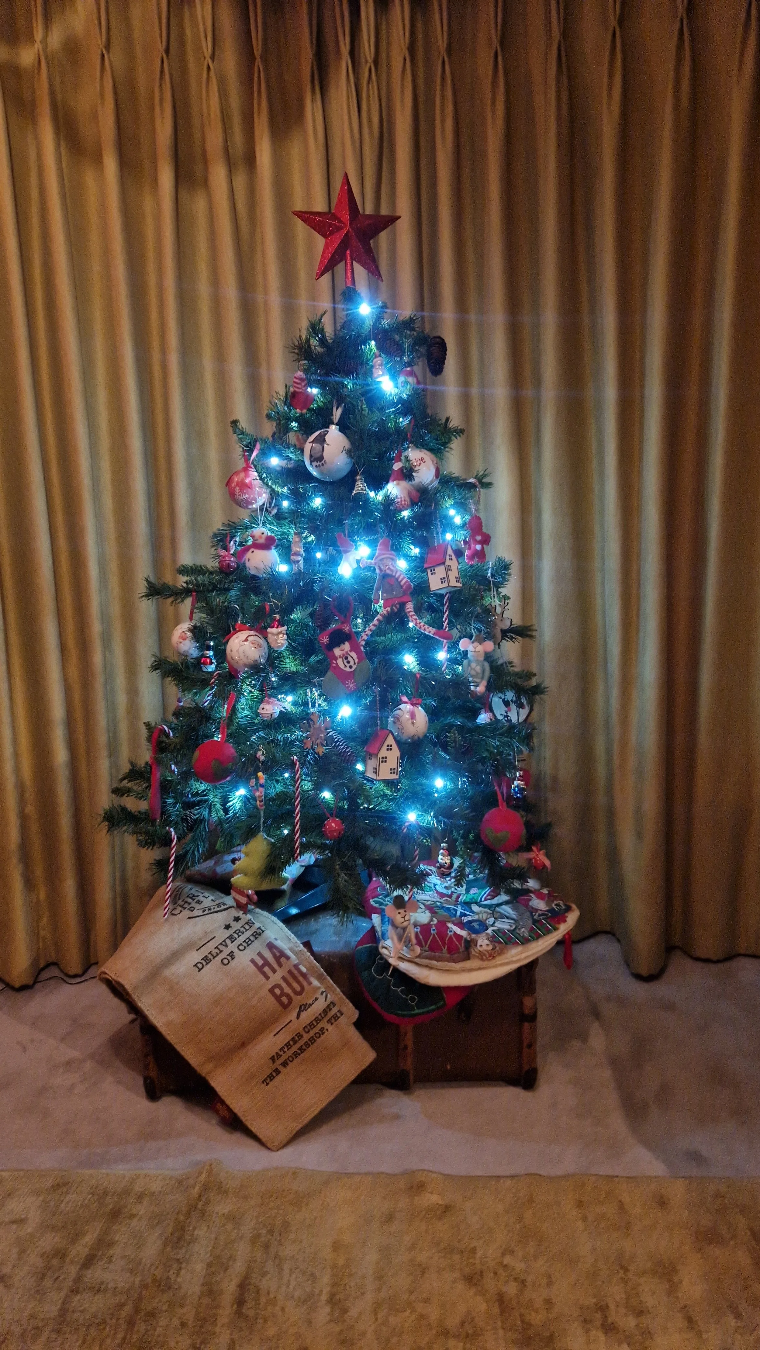 A christmas tree showing all the kids decorations, twinkly lights in an living room with full length curtains. created by Studio Dee interior design.