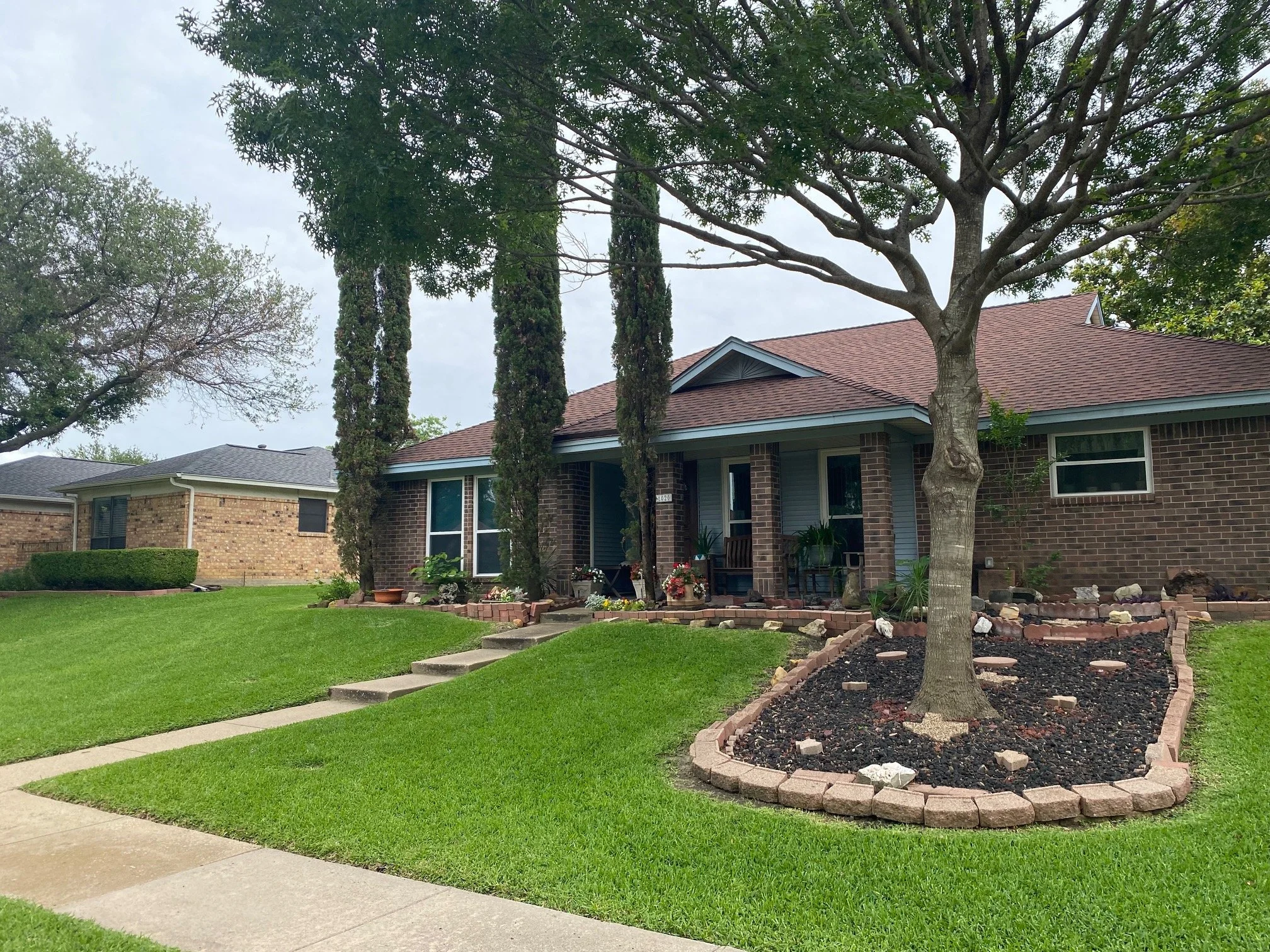 Front yard of a brick house with a large tree and a landscaped garden bed.