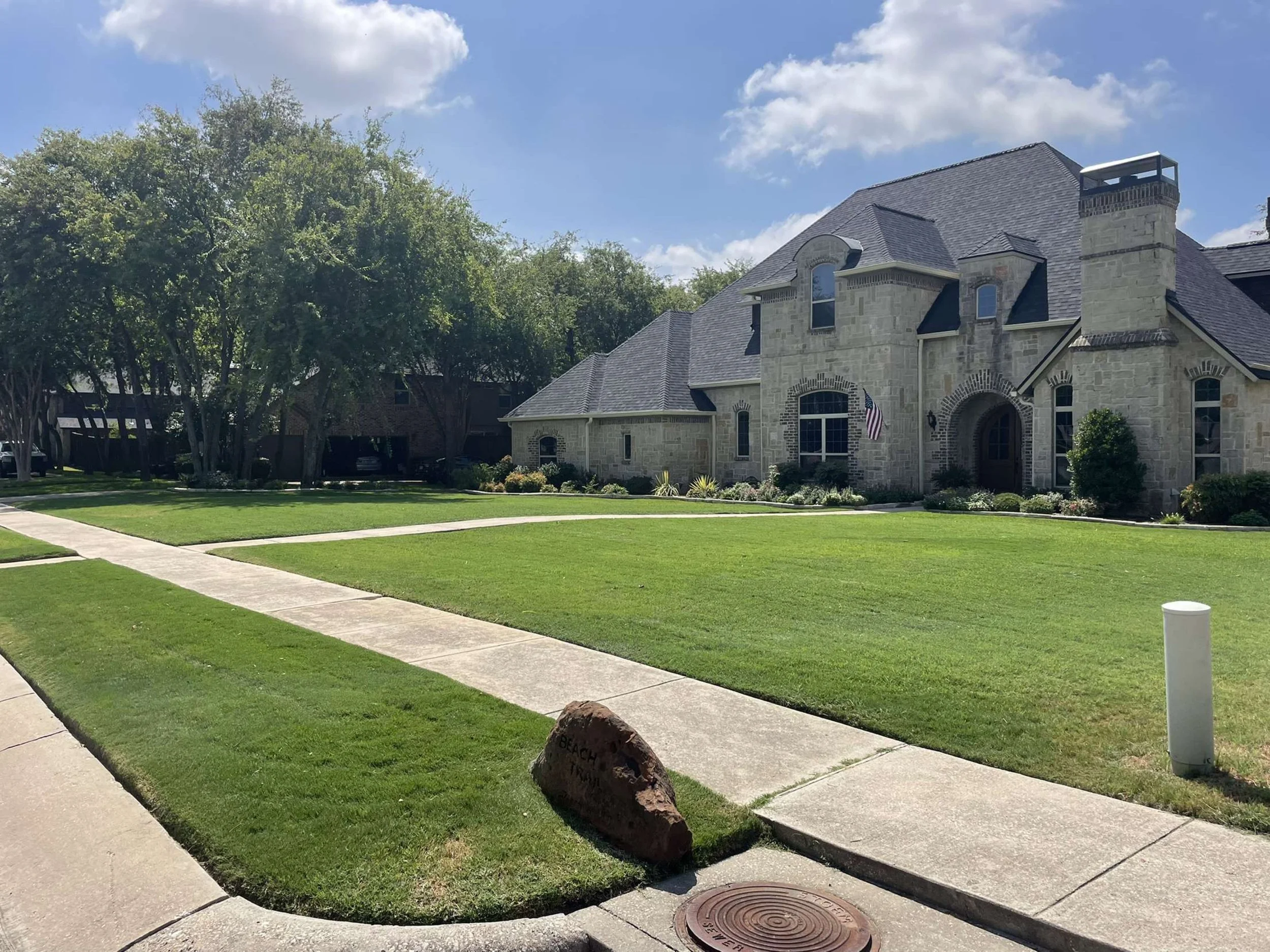 A large stone house with a well-manicured lawn, sidewalk, trees, and a clear blue sky.