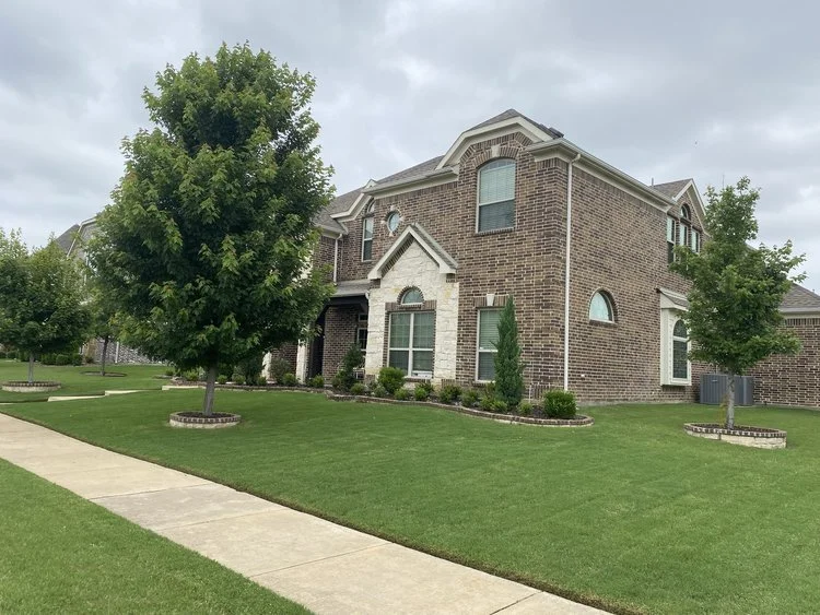 A two-story brick house with arched windows and a front porch, surrounded by a well-maintained lawn and trees, on a cloudy day.