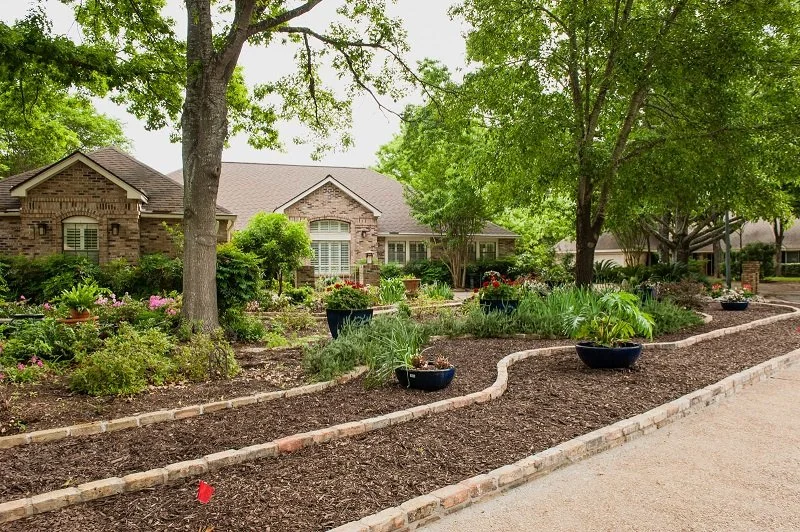 A landscaped front yard with trees, bushes, and flowers in pots and garden beds in front of a brick house with a gabled roof.