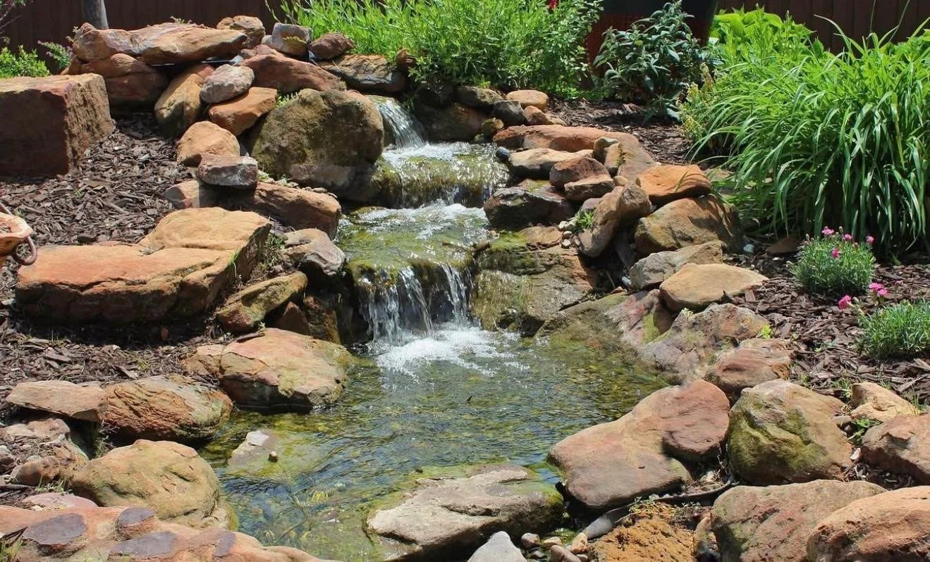 A small garden pond with a waterfall feature, surrounded by rocks and greenery, including various plants and flowers.