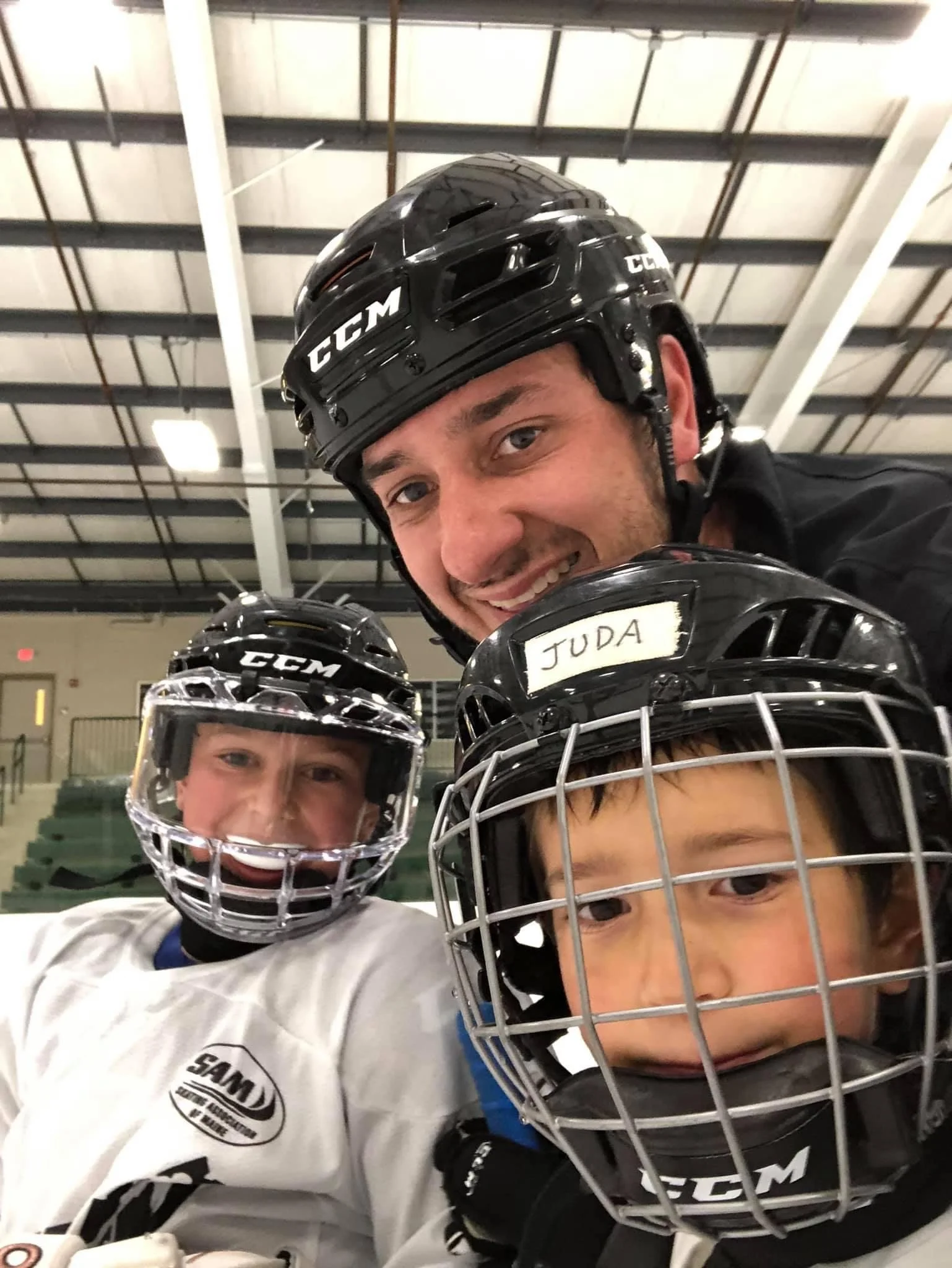 A man and two children wearing hockey gear, including helmets and jerseys, taking a selfie inside an ice rink.