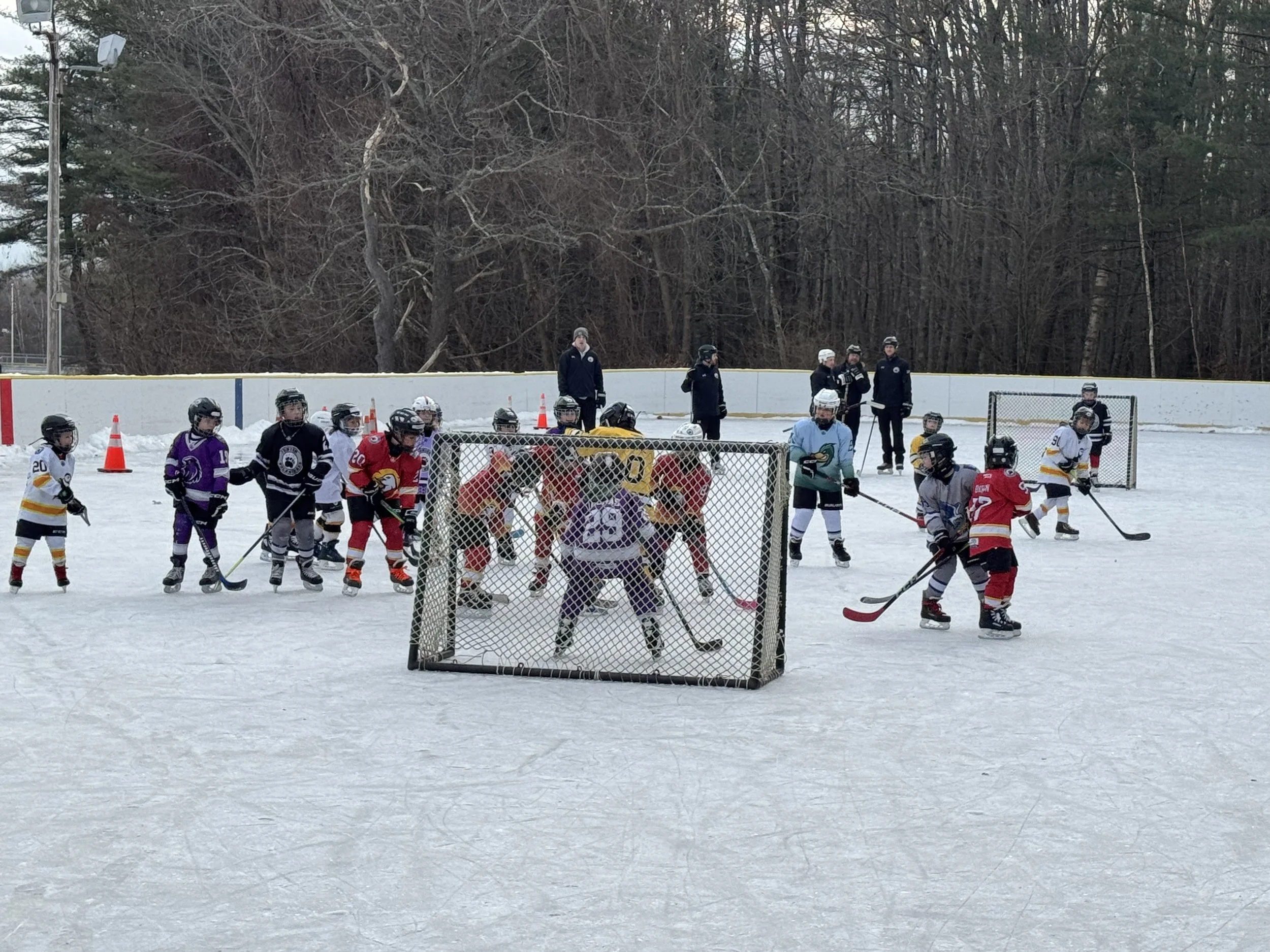 Children playing ice hockey on an outdoor rink with coaches and spectators in the background, surrounded by a chain-link goal.
