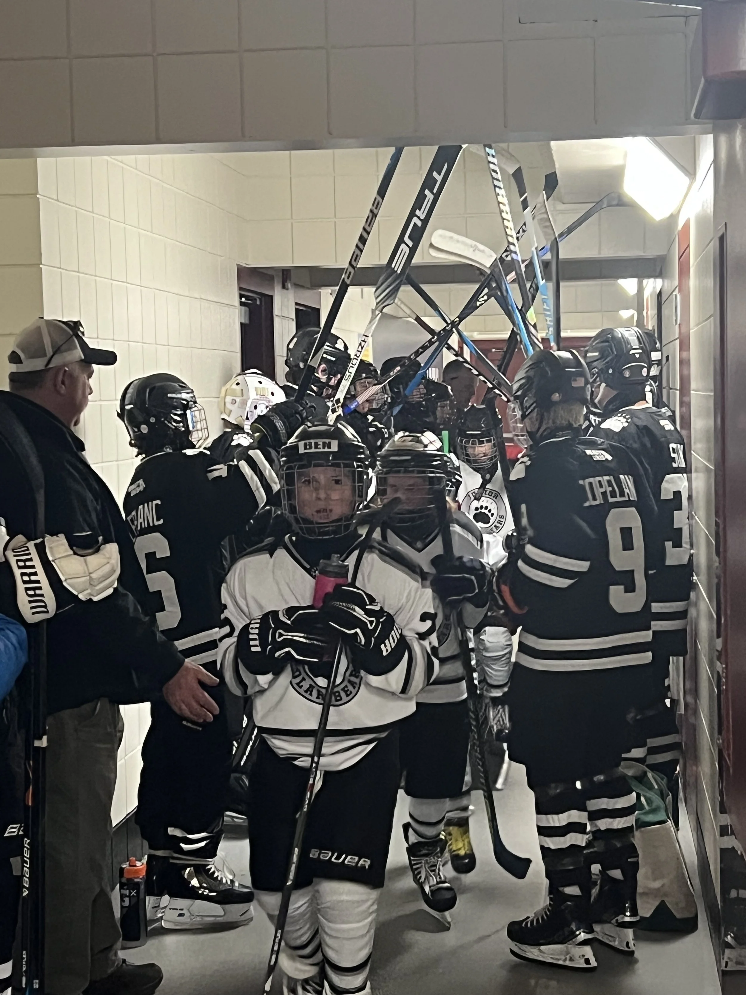 Hockey players in black and white uniforms with helmets and gloves, standing in a hallway before a game, some holding hockey sticks, with a coach or staff member to the left.