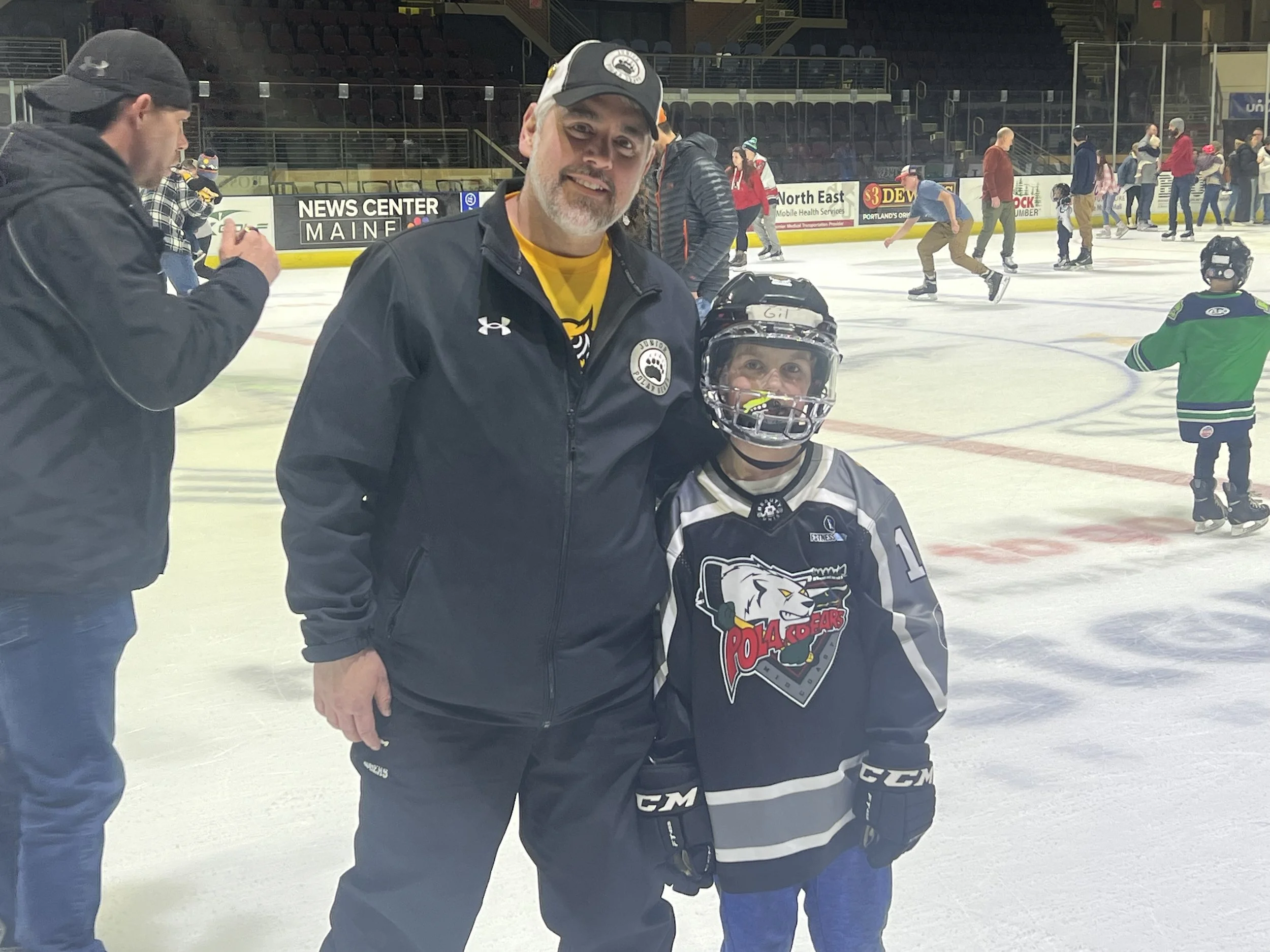 A man and a young hockey player standing on an ice rink, smiling for the photo, with other people and kids skating in the background.