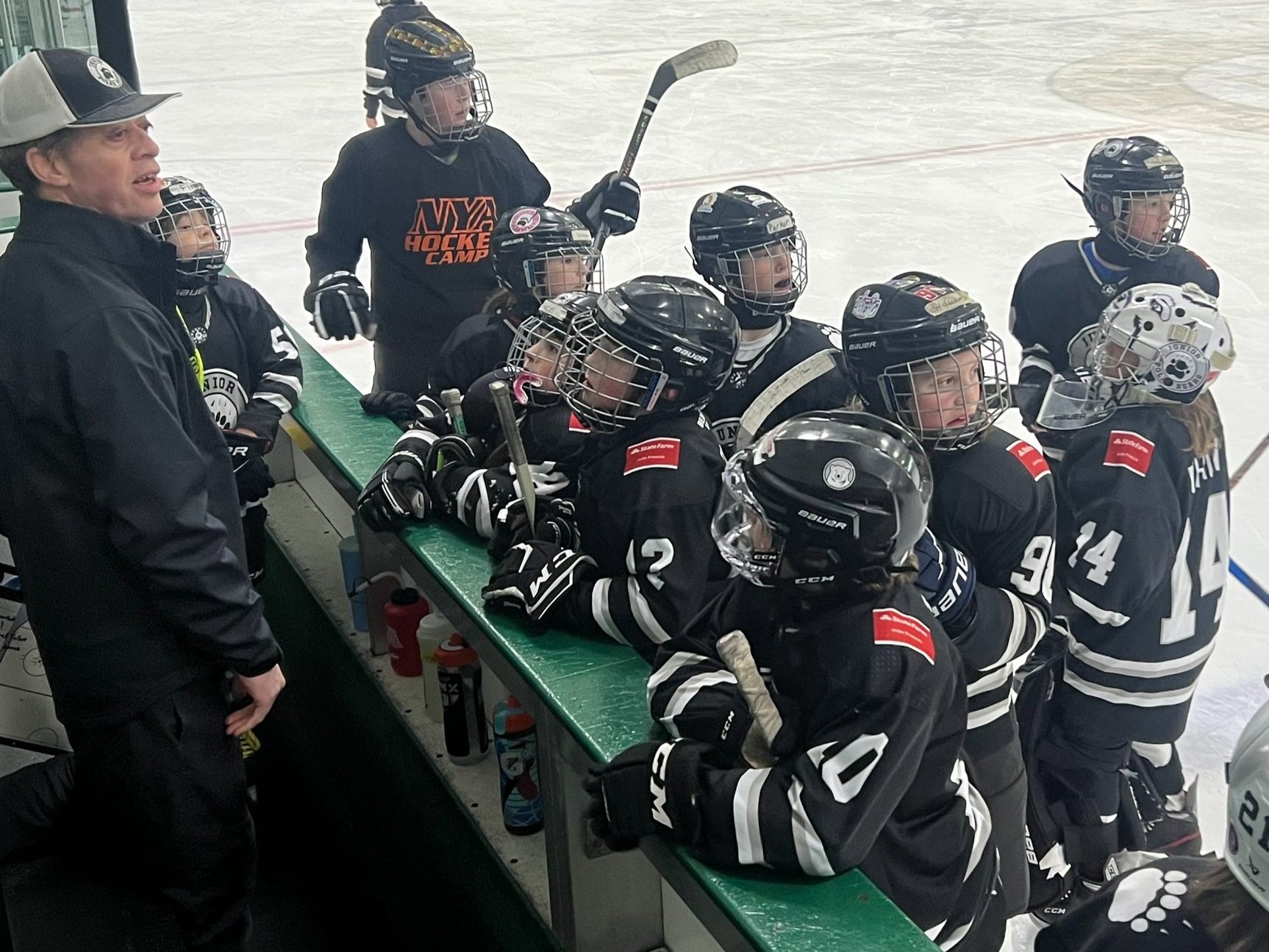 A youth hockey coach speaking to a team of young hockey players gathered at the bench during a game or practice on an ice rink.