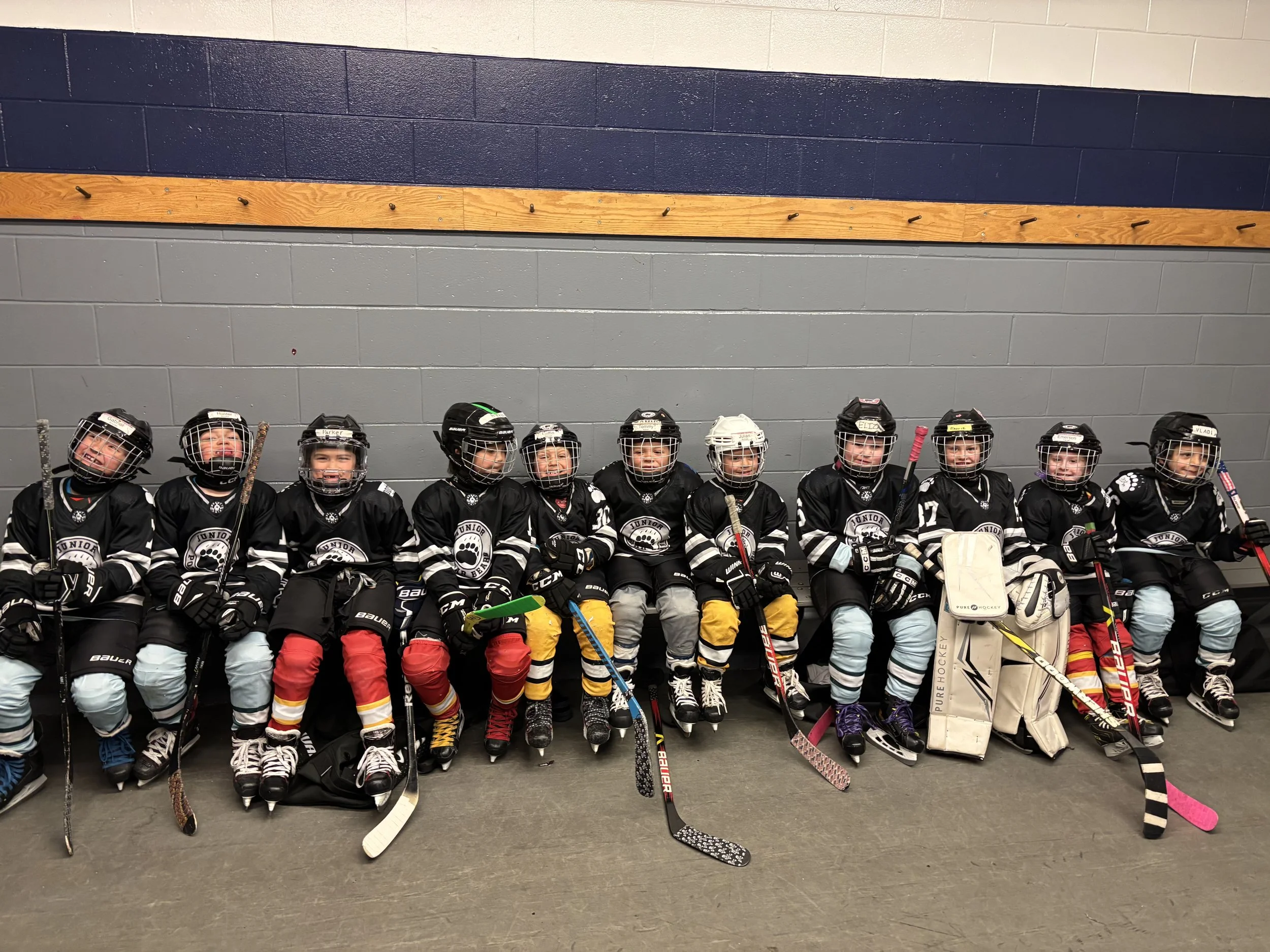 A group of young children in ice hockey uniforms sitting on a bench in an indoor rink, wearing helmets, gloves, and skates.