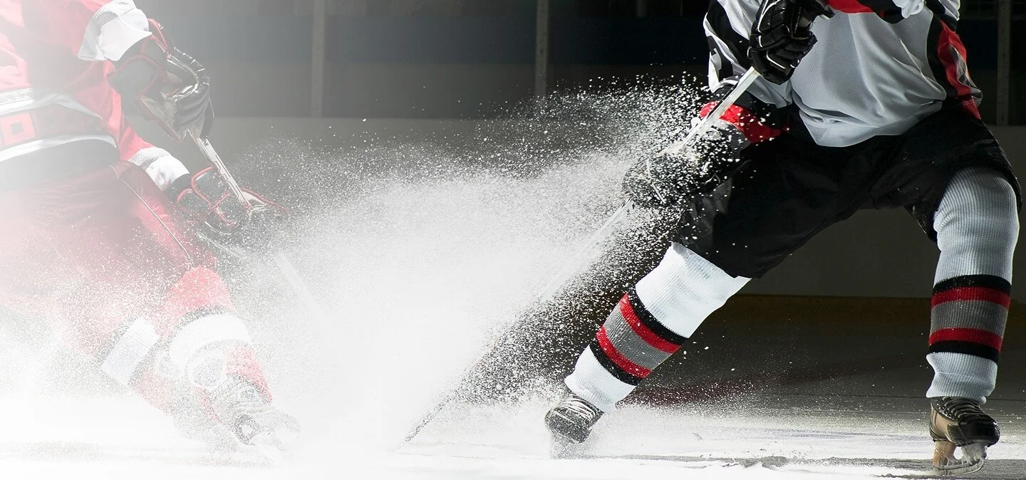 Hockey players in a game on the ice, with one player falling or being checked, and snow spray from the ice.