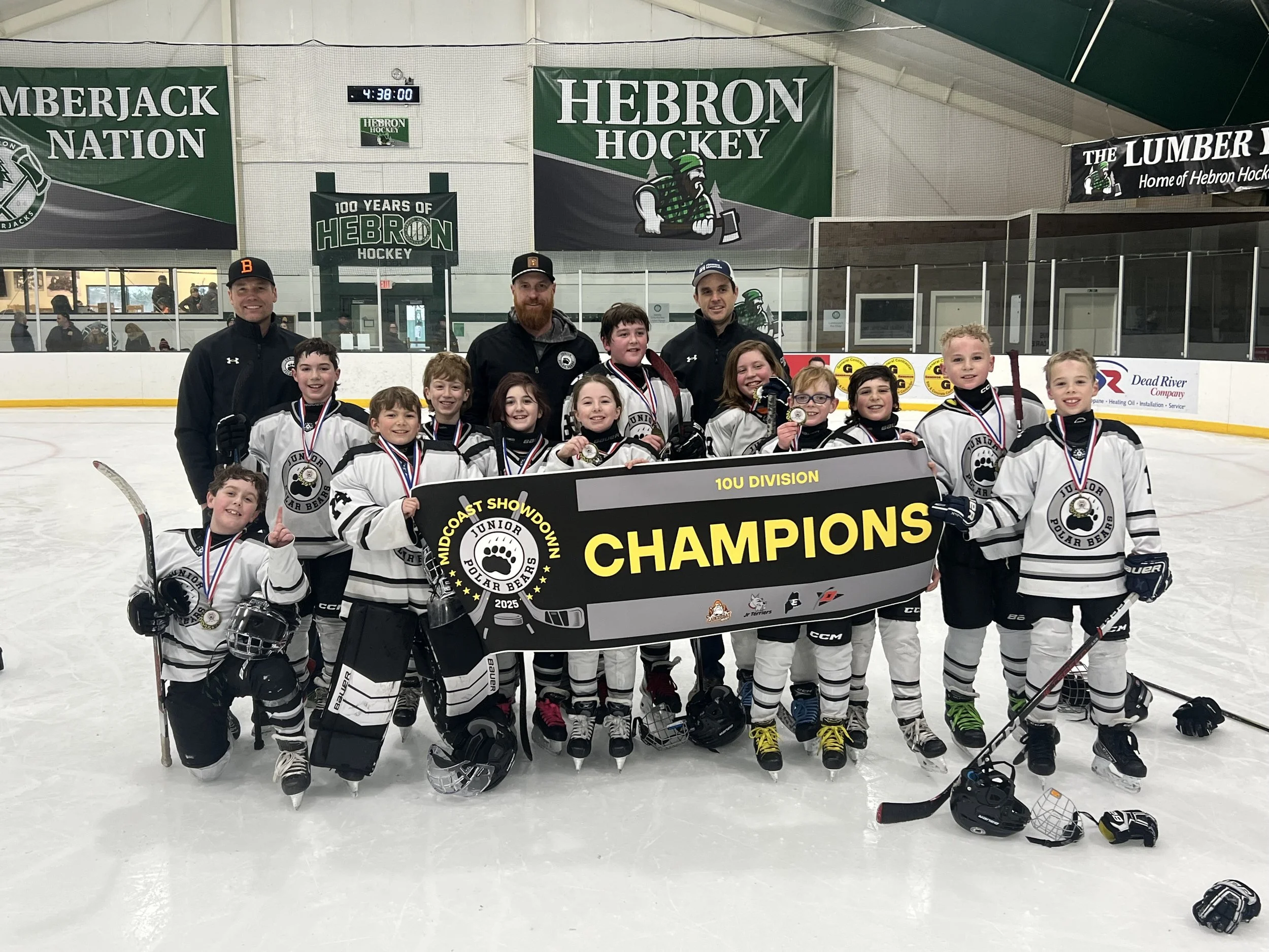 Youth hockey team and coaches celebrating their championship win on an ice rink, holding a large black and yellow banner that reads 'CHAMPIONS'. The team wears white jerseys with black accents, and medals around their necks.