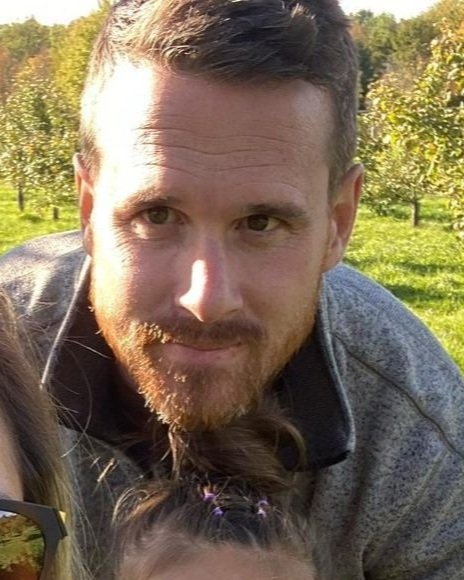 A man with short brown hair and a beard outdoors in a sunny orchard with green trees.