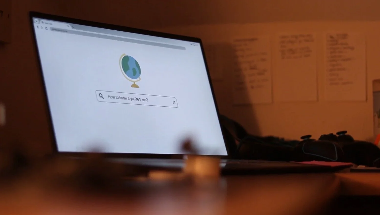 Laptop screen displaying Google search page with a globe icon and query 'How to know if you're trans?'. Office desk with a game controller and papers in the background.